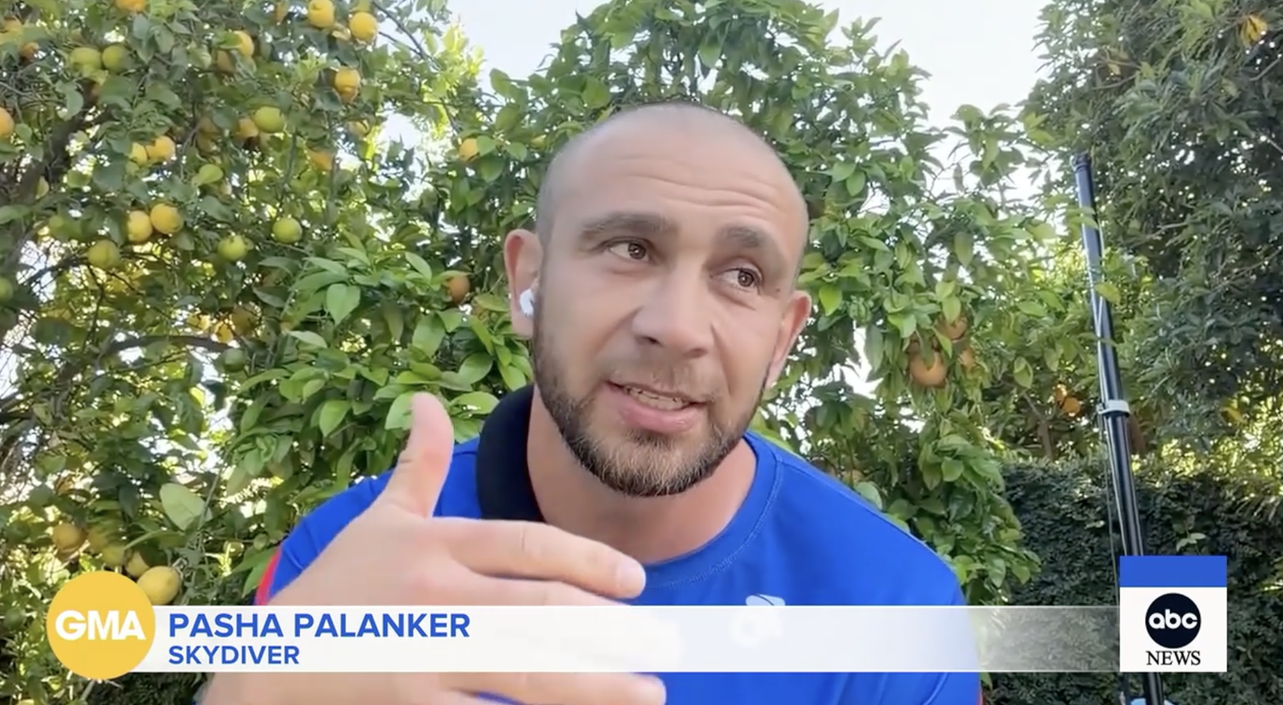 A man with a beard and earbuds, wearing a blue shirt, appears to be speaking while gesturing with his hand in an outdoor setting with fruit trees and greenery in the background during a TV interview.