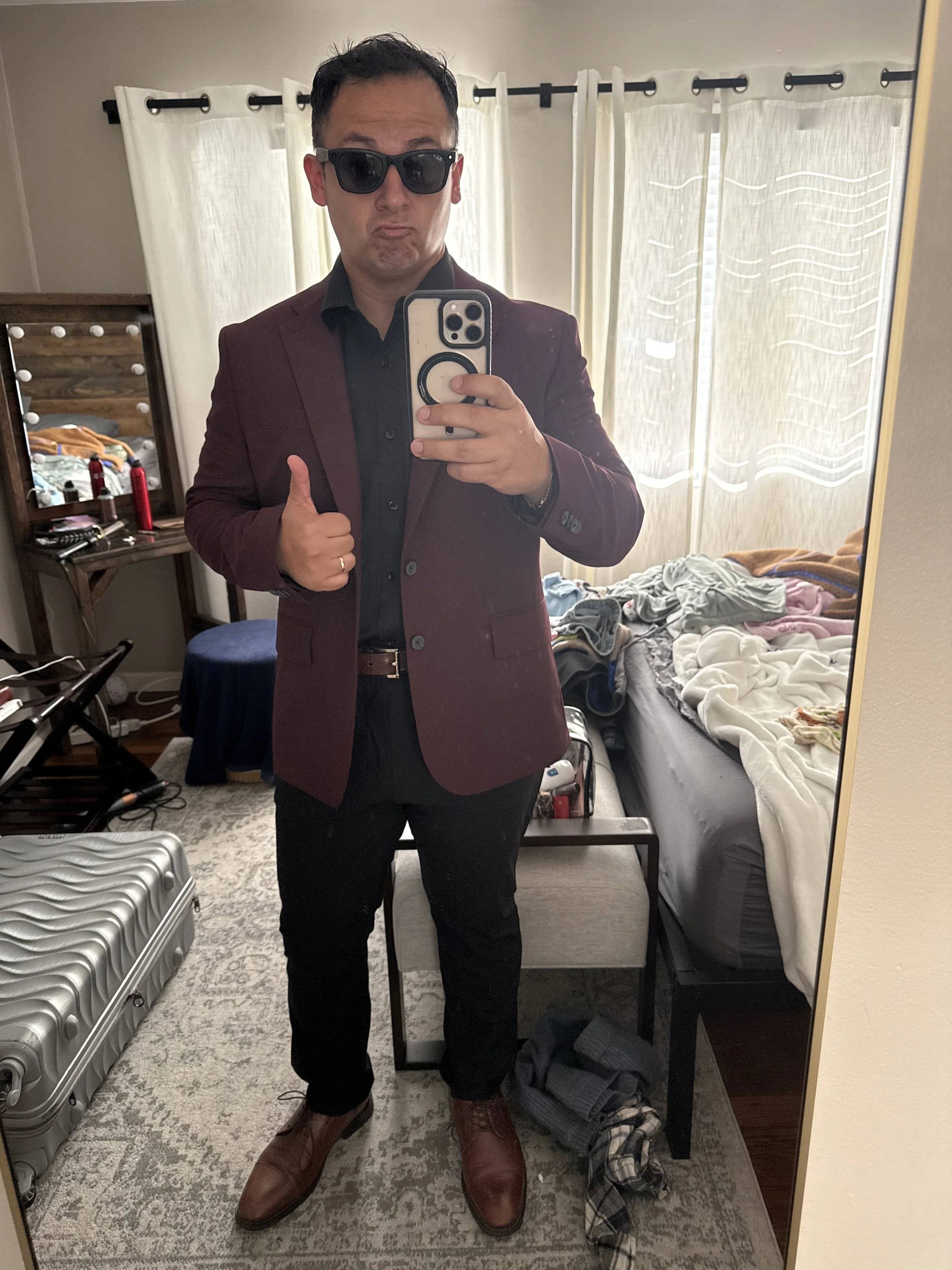 A man taking a mirror selfie in a bedroom, wearing sunglasses, a dark shirt, a maroon blazer, black pants, and brown boots, with messy clothes on the bed and cluttered furniture in the background.