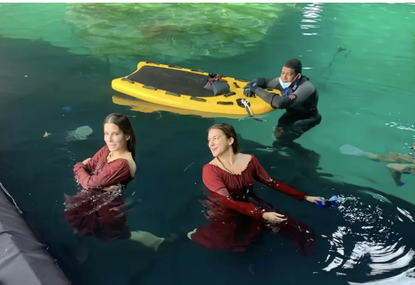 Two women in red dresses are in a body of water, smiling and posing for a photo, with one woman in the foreground and the other slightly behind her. A man in black waterproof gear and a face mask is holding onto a yellow rescue board in the backgroun