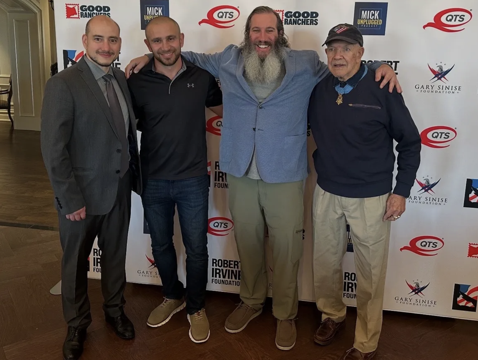 Five men standing together at a promotional event with a backdrop featuring logos such as QTS, Good Ranchers, Mick Unplugged, and Gary Sinise Foundation. They are smiling, with four of them having their arms around each other's shoulders.