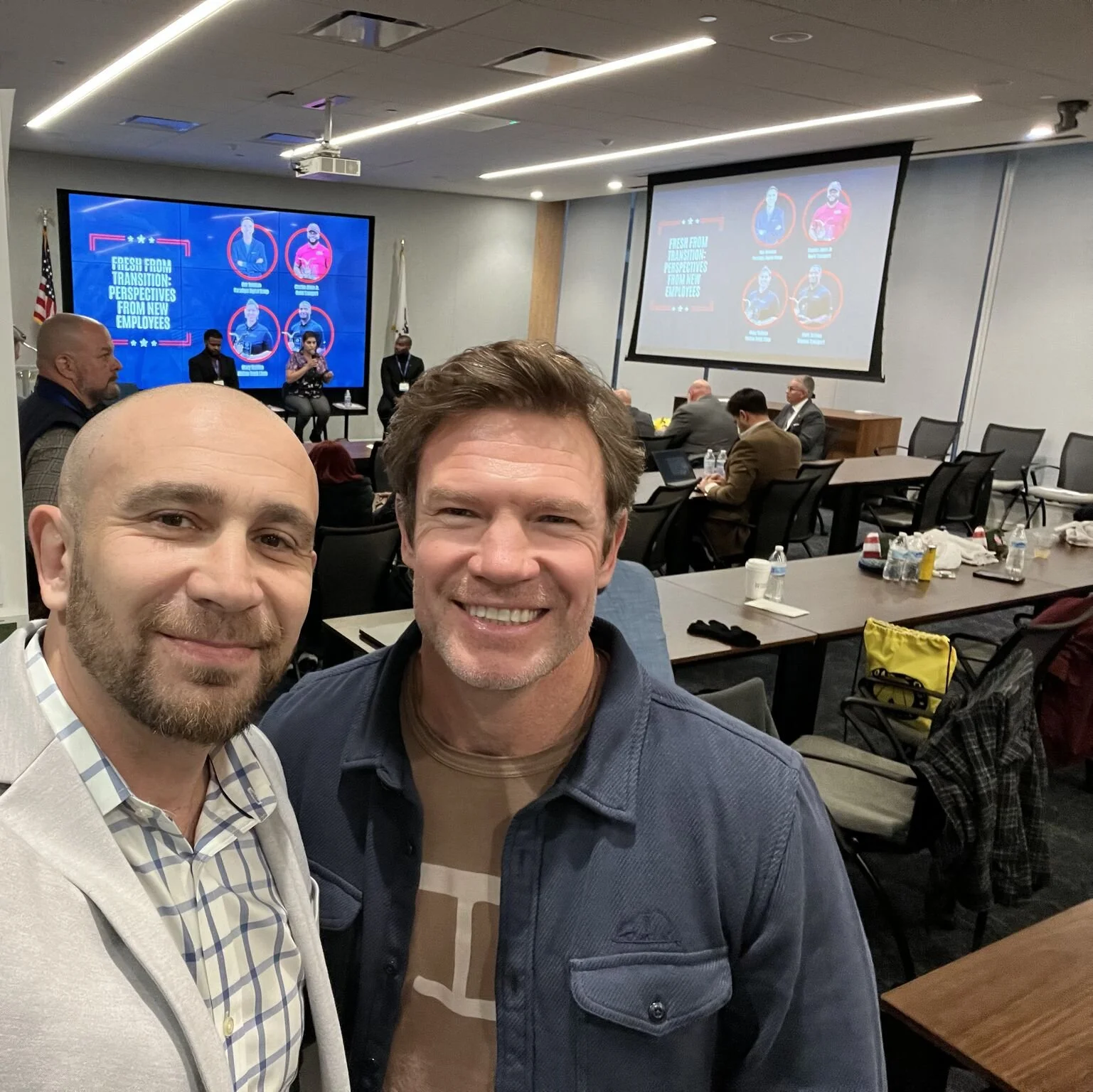 Two men taking a selfie at a conference or seminar, with a presentation screen behind them showing speakers and a slide about onboarding new employees. Other attendees are seated and engaged in discussion or working.