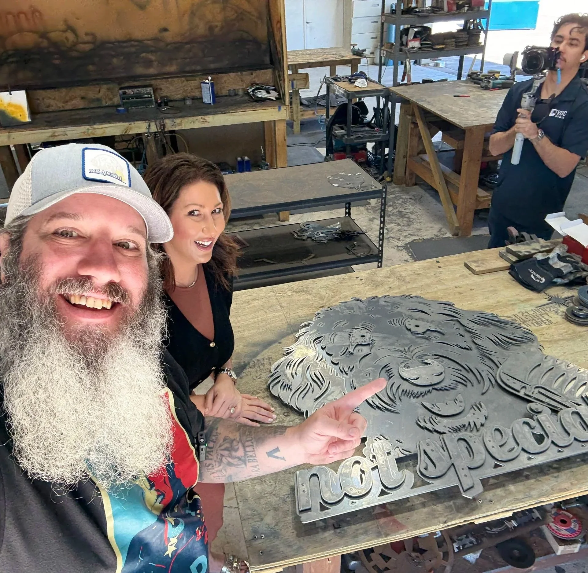 Man with a long beard, wearing a hat and colorful shirt, smiling at camera while pointing at a metal cutout with various shapes on a workbench. Two women are also partly visible observing. The background shows a workshop with tools and shelves.