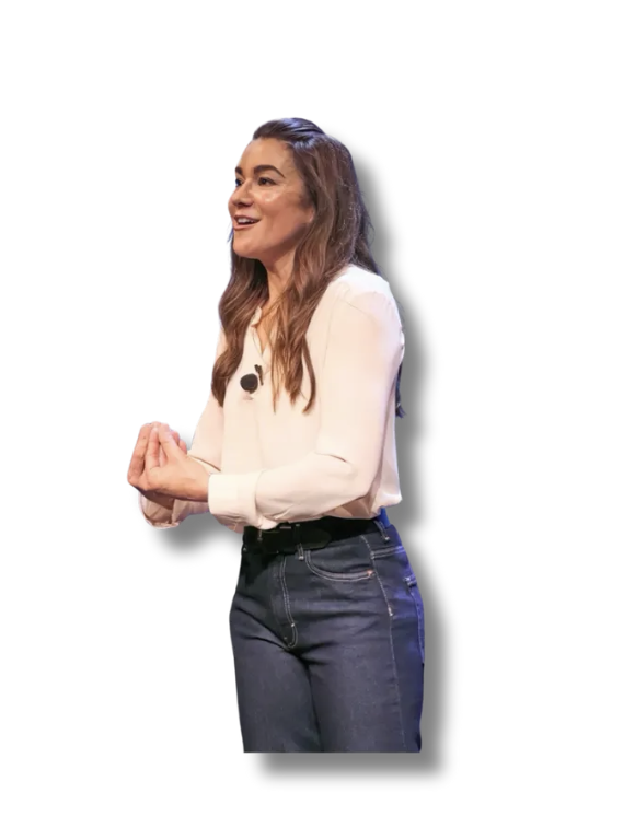 A woman with long brown hair wearing a white blouse and dark jeans, smiling and gesturing with her hands during a presentation.