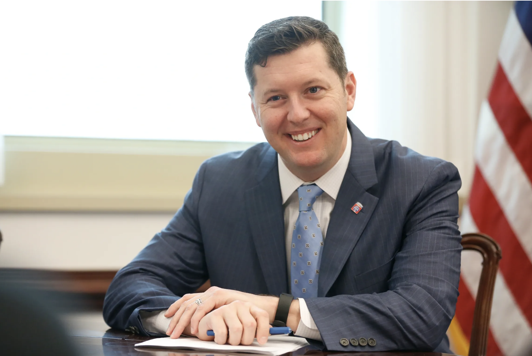 A smiling man in a blue checkered suit with a light blue tie, sitting at a table in a room with large windows and an American flag in the background.
