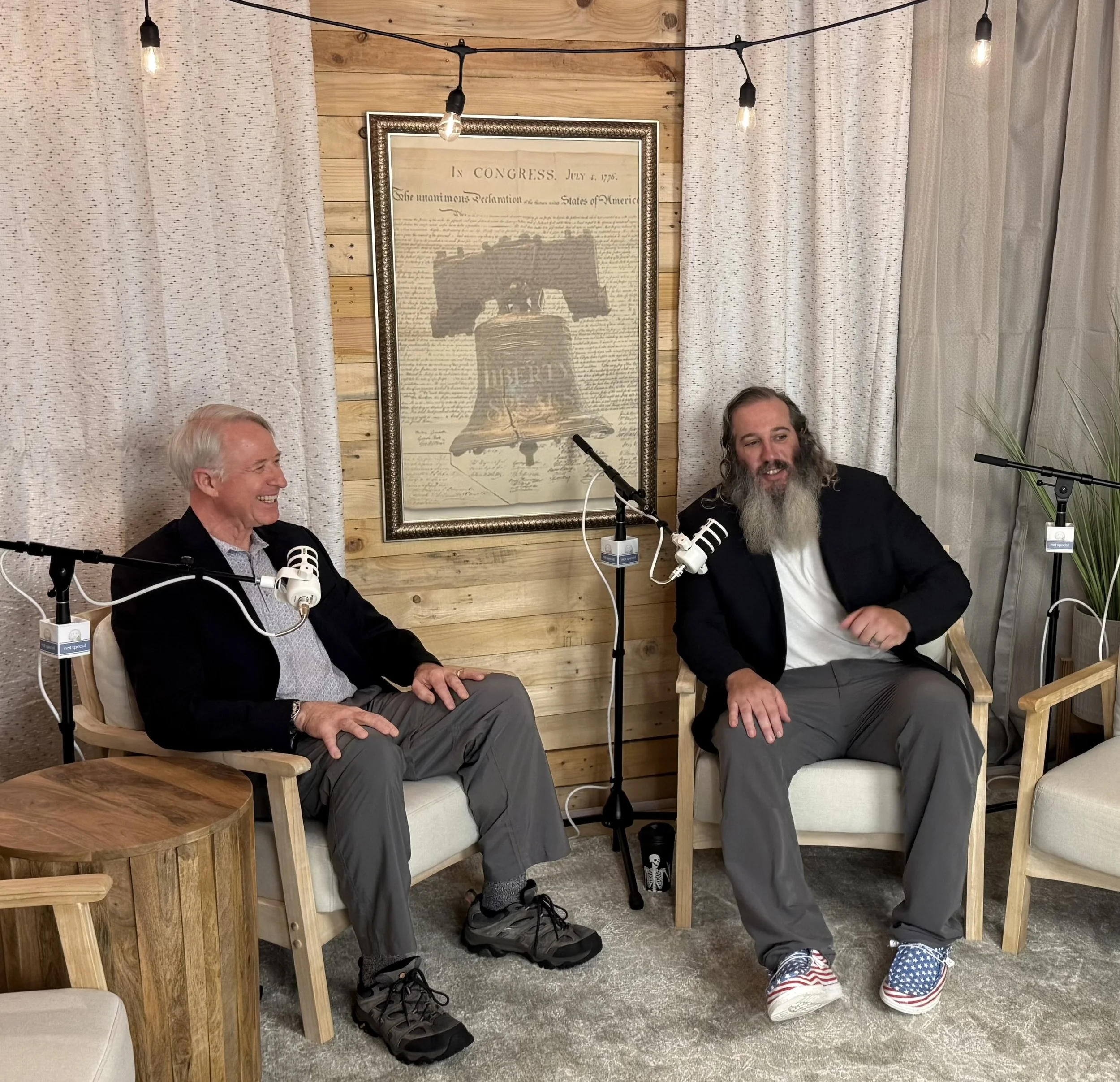 Two men are sitting and talking in a room decorated with wooden walls and curtains. There are microphones in front of them, suggesting a recording or interview setting. The man on the left has white hair and is smiling, wearing a black jacket and a s