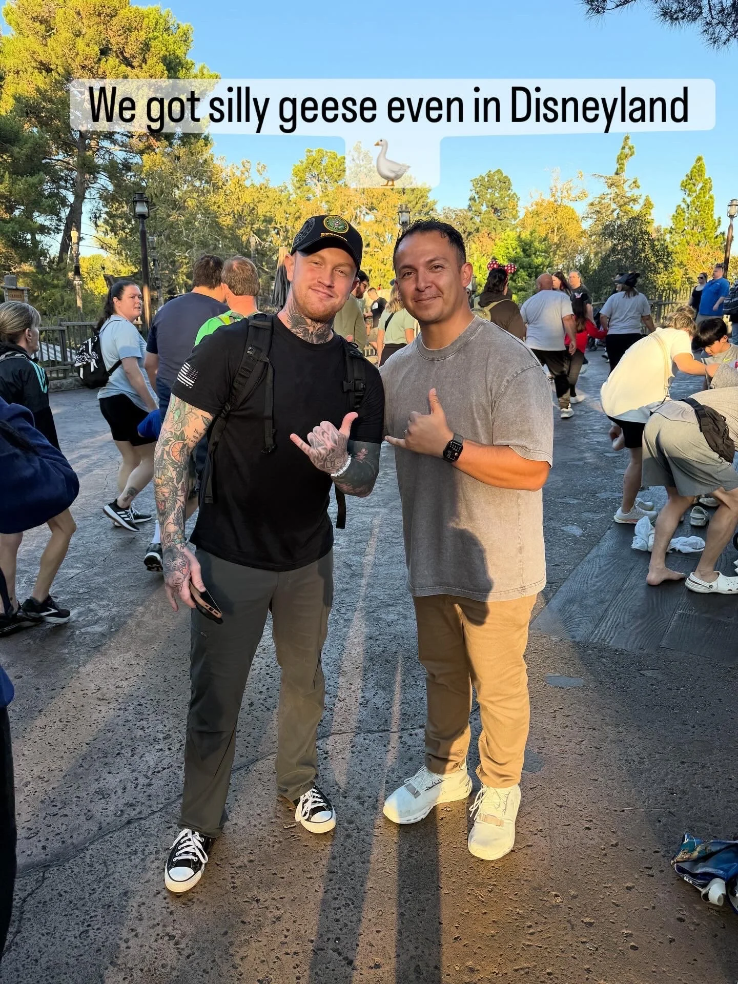 Two young men standing close together in an amusement park, smiling and making the shaka sign with one hand each, surrounded by other visitors.