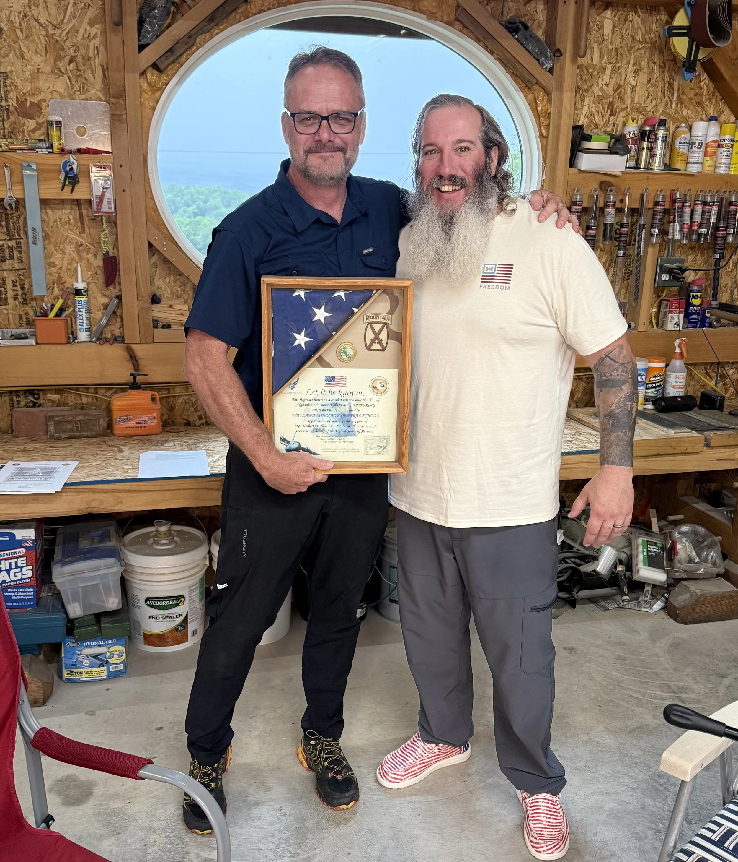 Two men standing in a wood workshop, smiling, one holding a framed display with American flag and military insignia, and the other dressed casually, both wearing colorful sneakers, with various tools and supplies on shelves behind them and a round wi