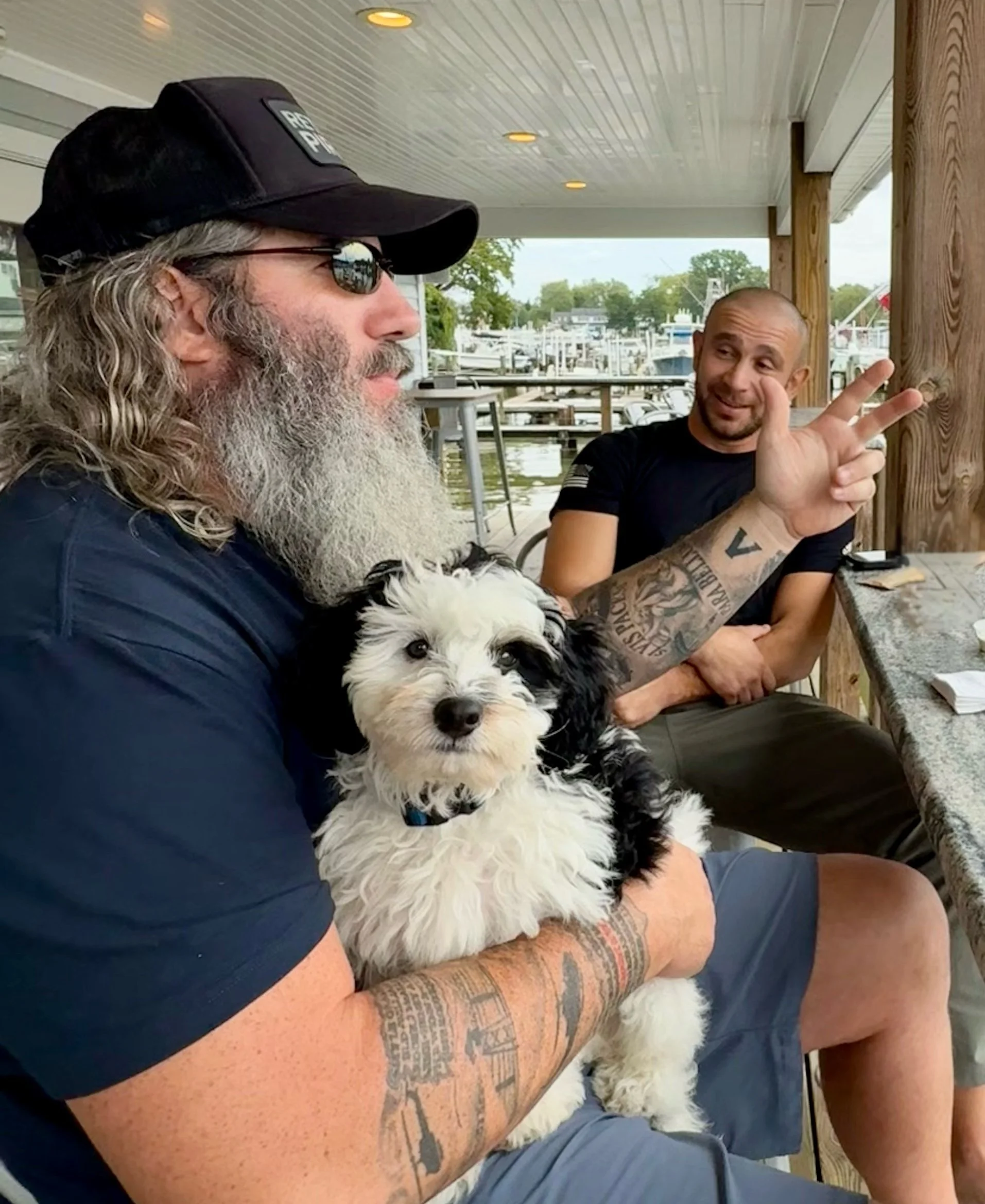 Two men are sitting at an outdoor table, engaging in conversation. The man on the left is wearing sunglasses, a black cap, and has a long gray beard. He has tattoos on his arm and is holding a black and white dog on his lap. The man on the right, who