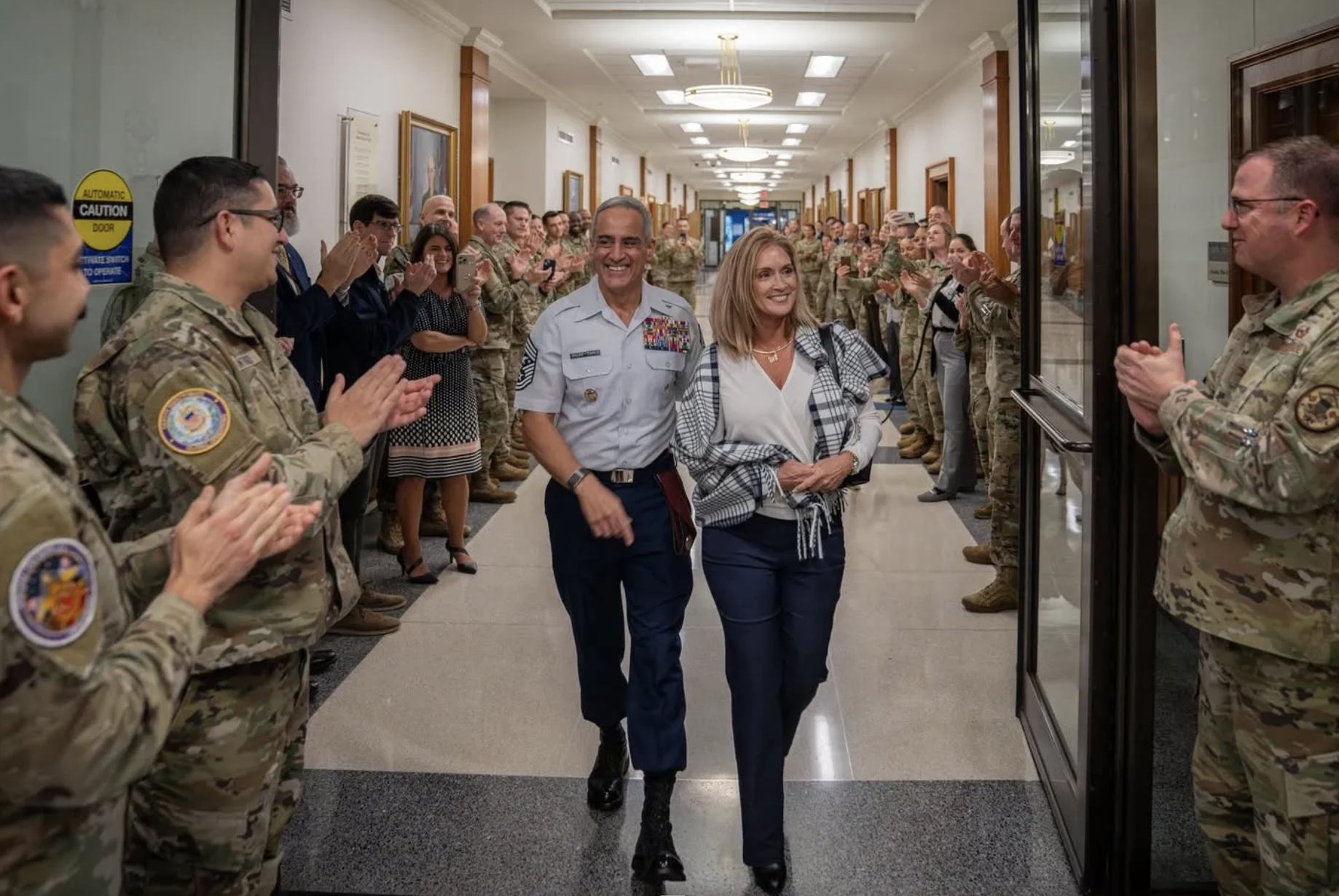 A group of military personnel in uniform clapping and cheering as a man and woman walk down a hallway, smiling. The man appears to be a military officer, and the woman is wearing a checkered shirt and neckless. The hallway is decorated with framed po