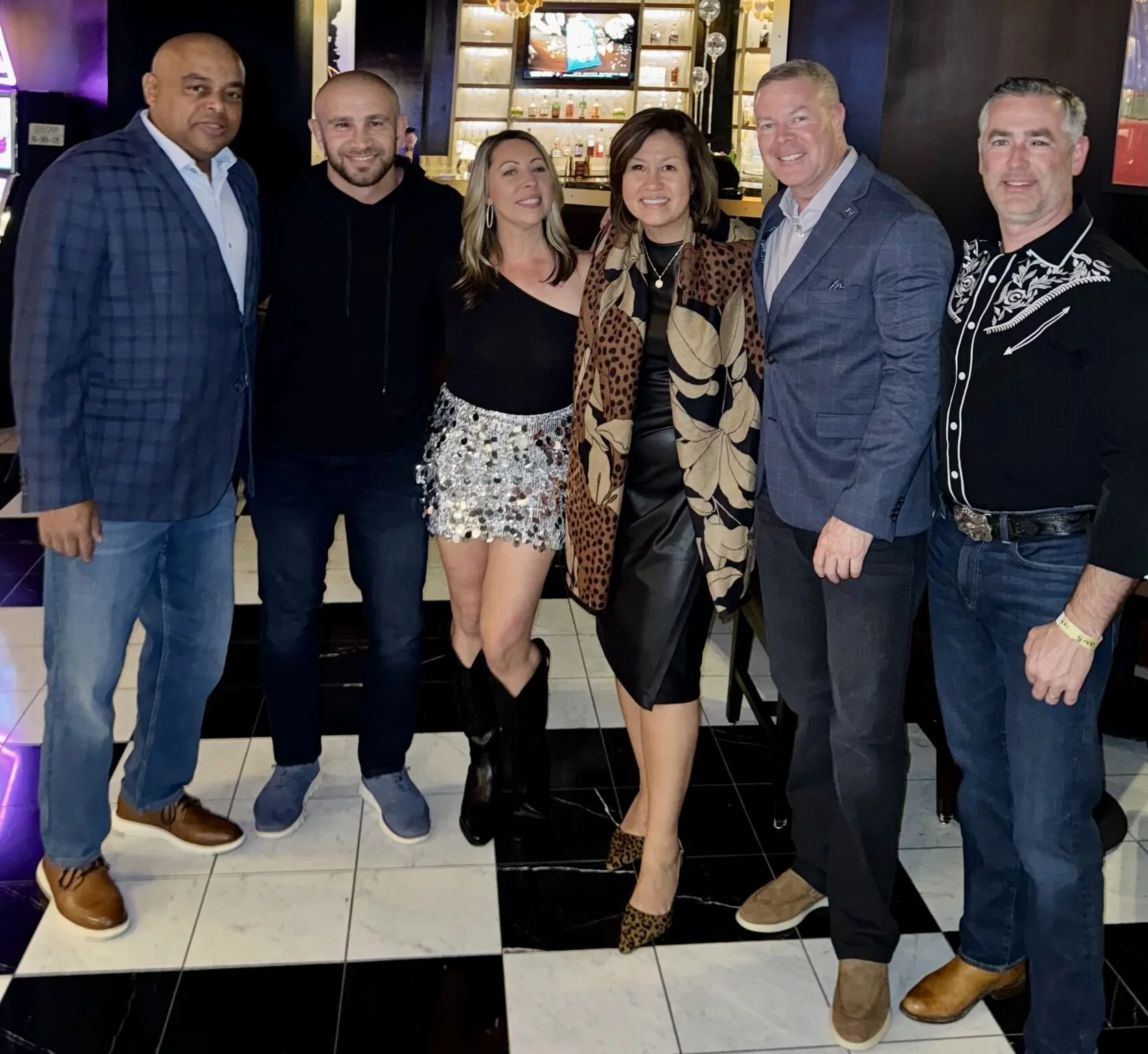 A group of seven diverse adults standing together in a bar or restaurant, smiling for the photo. There are three women and four men, with a bar displaying bottles and a television screen in the background.