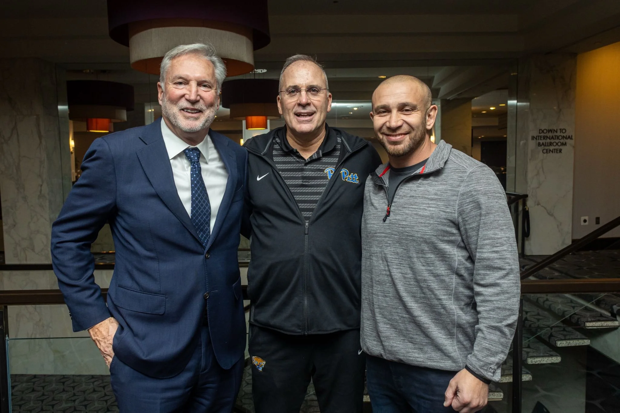 Three men standing together indoors, smiling for a photo. The man on the left is wearing a blue suit with a white shirt and blue tie. The man in the middle is wearing glasses and a black jacket with a sports logo. The man on the right is wearing a gr