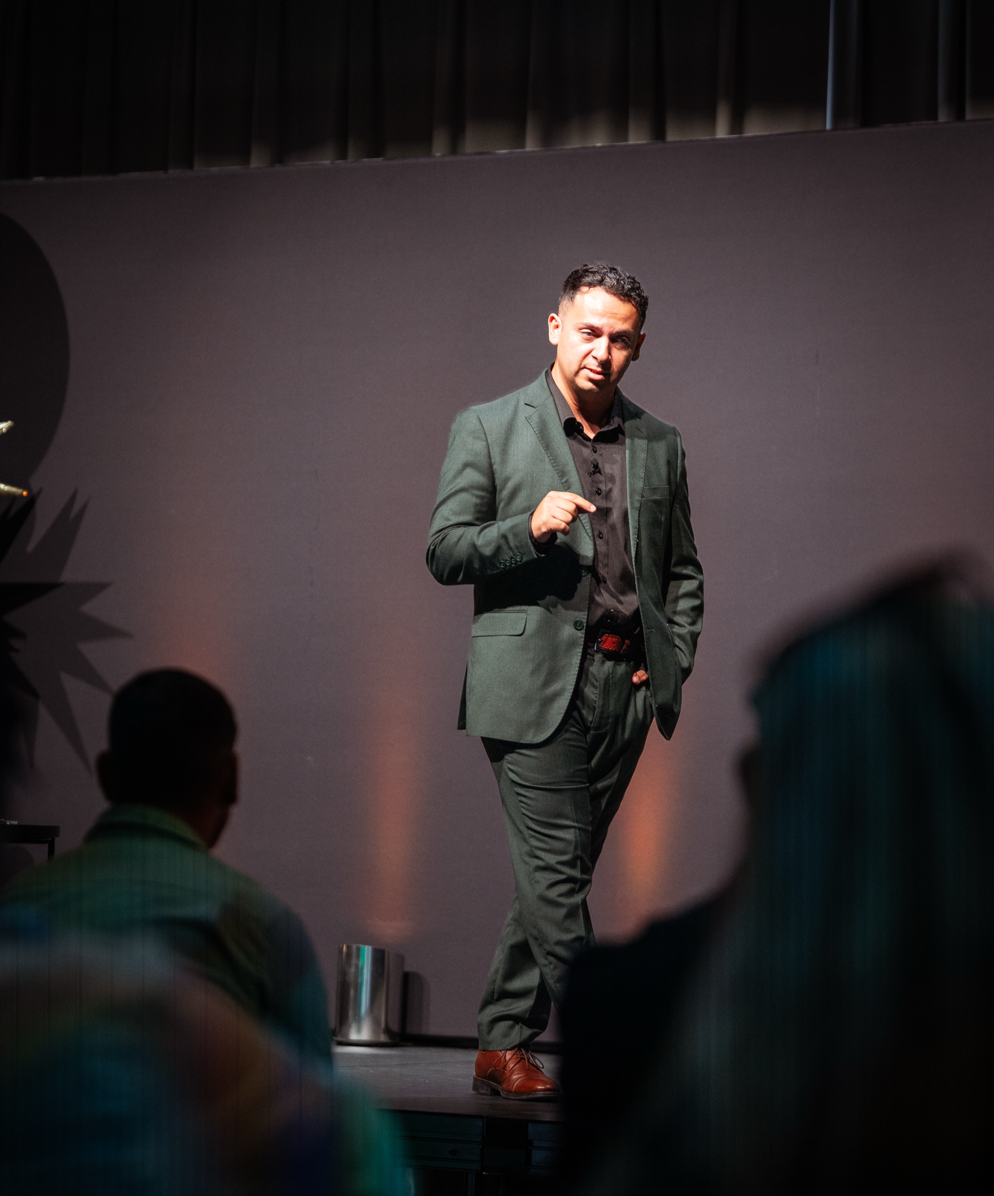 A man in a dark green blazer and black shirt speaking on stage with a dark background, while audience members listen.
