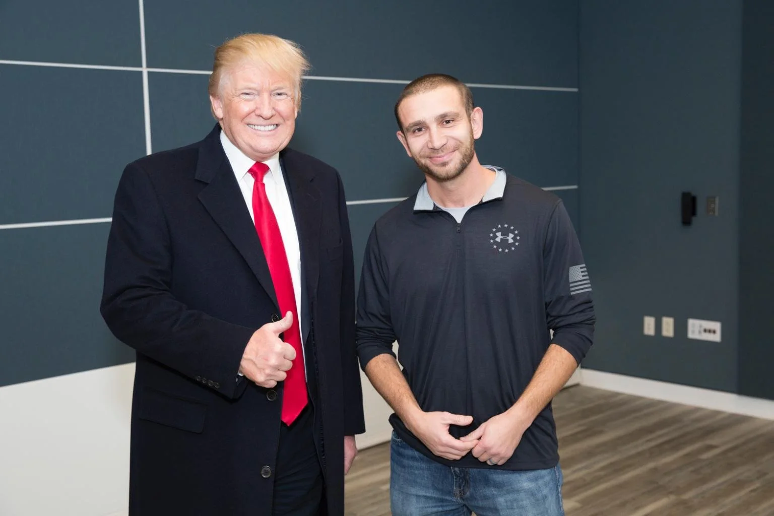Two men standing indoors, smiling for the camera. The man on the left is wearing a dark suit, white shirt, and red tie. The man on the right is dressed casually in a black jacket with patches and blue jeans.