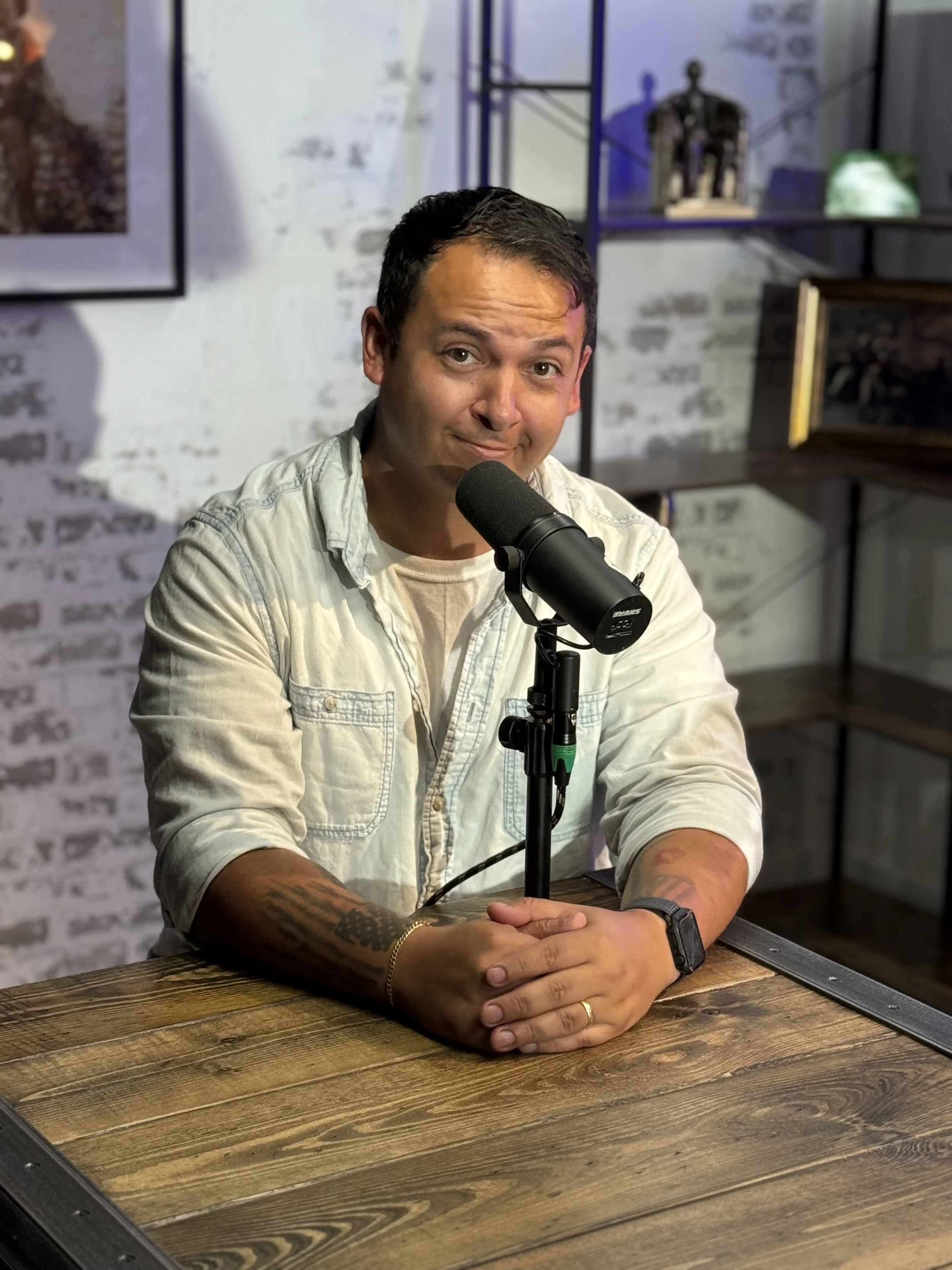 A man is sitting at a wooden table with his hands clasped in front of a microphone, wearing a light-colored denim shirt, with a brick wall and artwork behind him.
