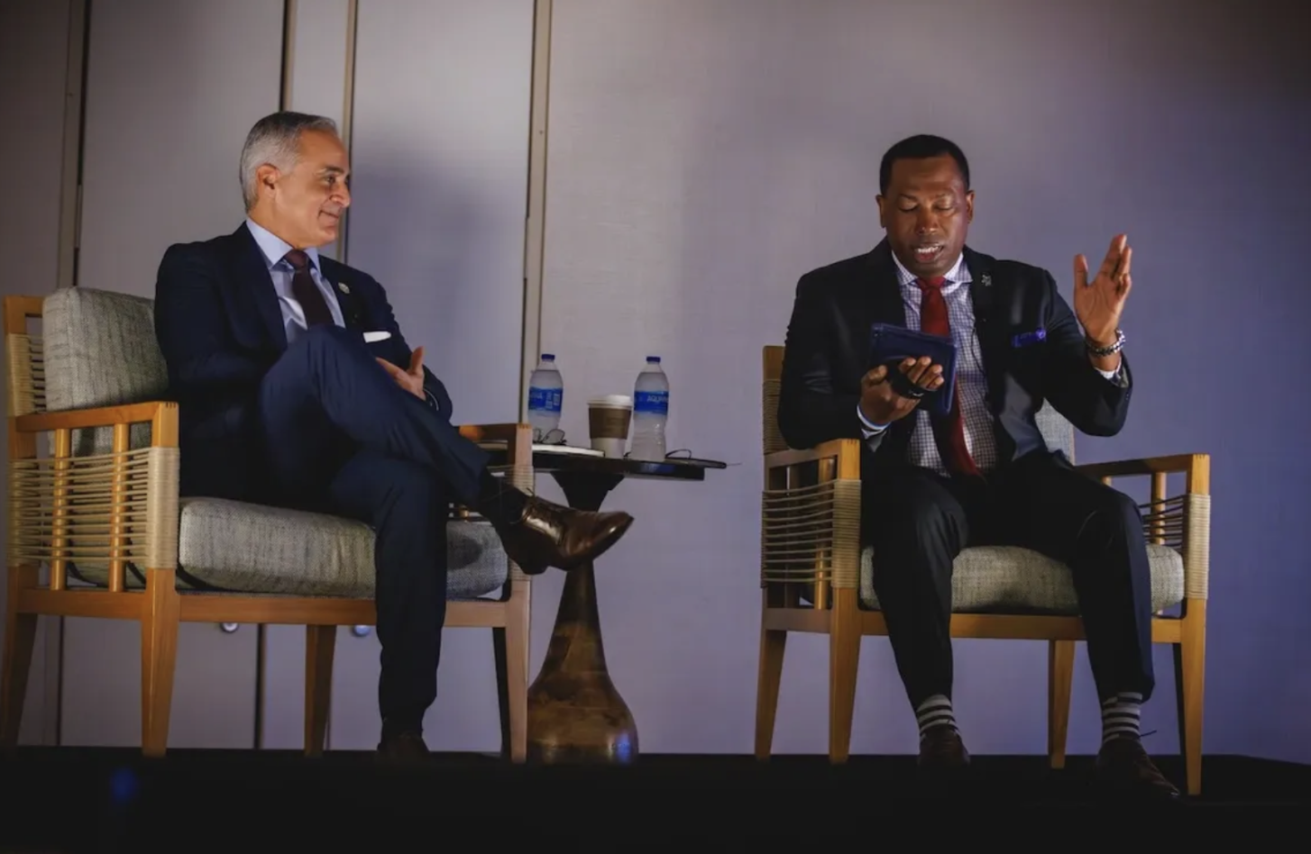 Two men in suits sitting on stage chairs during a panel discussion, with water bottles and cups on a side table between them.