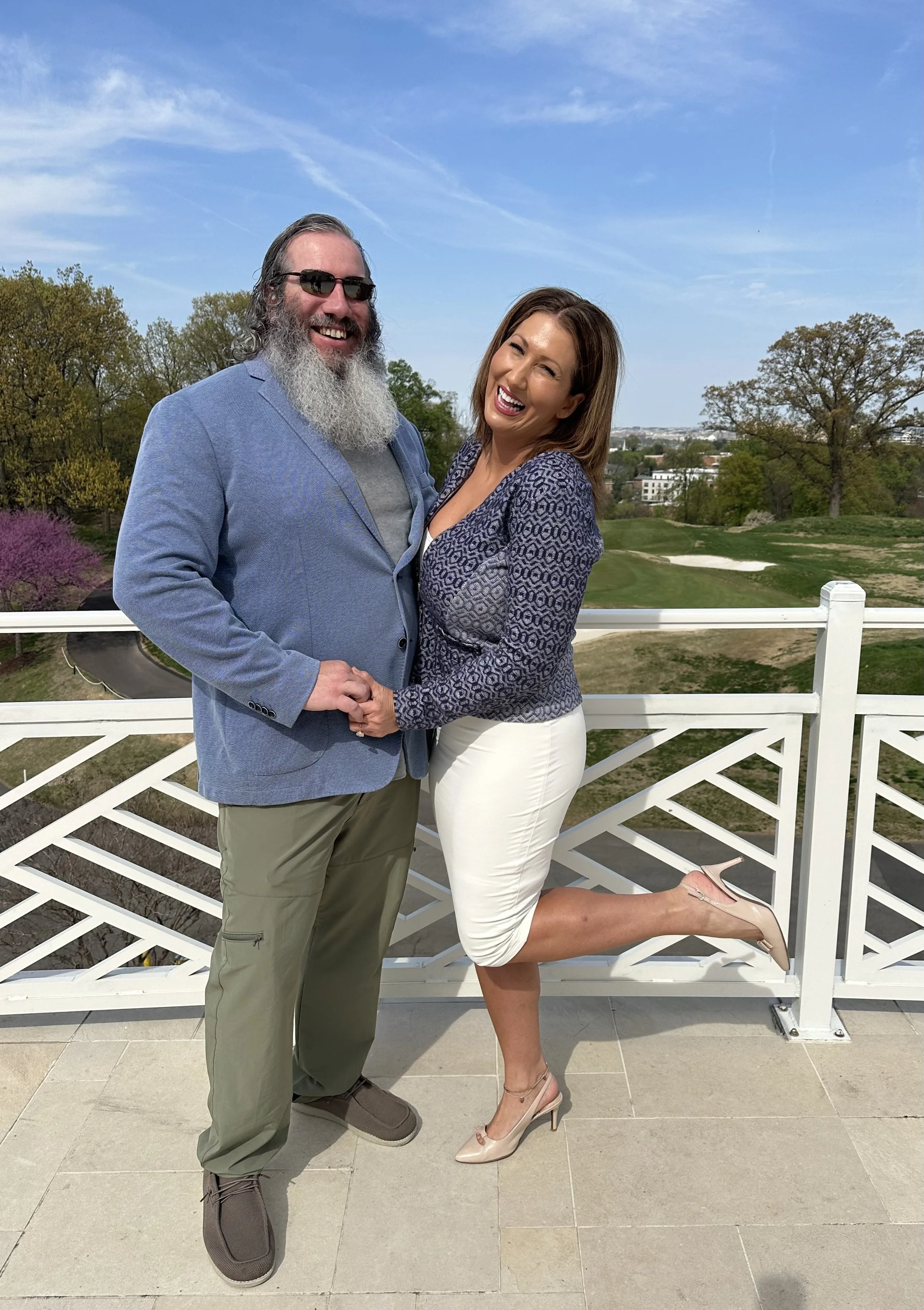A smiling man with a beard, wearing sunglasses, a light blue blazer, and khaki pants, stands next to a woman in a patterned blouse and white skirt. She is laughing, with one leg raised and her foot resting on a ledge, wearing nude high heels. They ar
