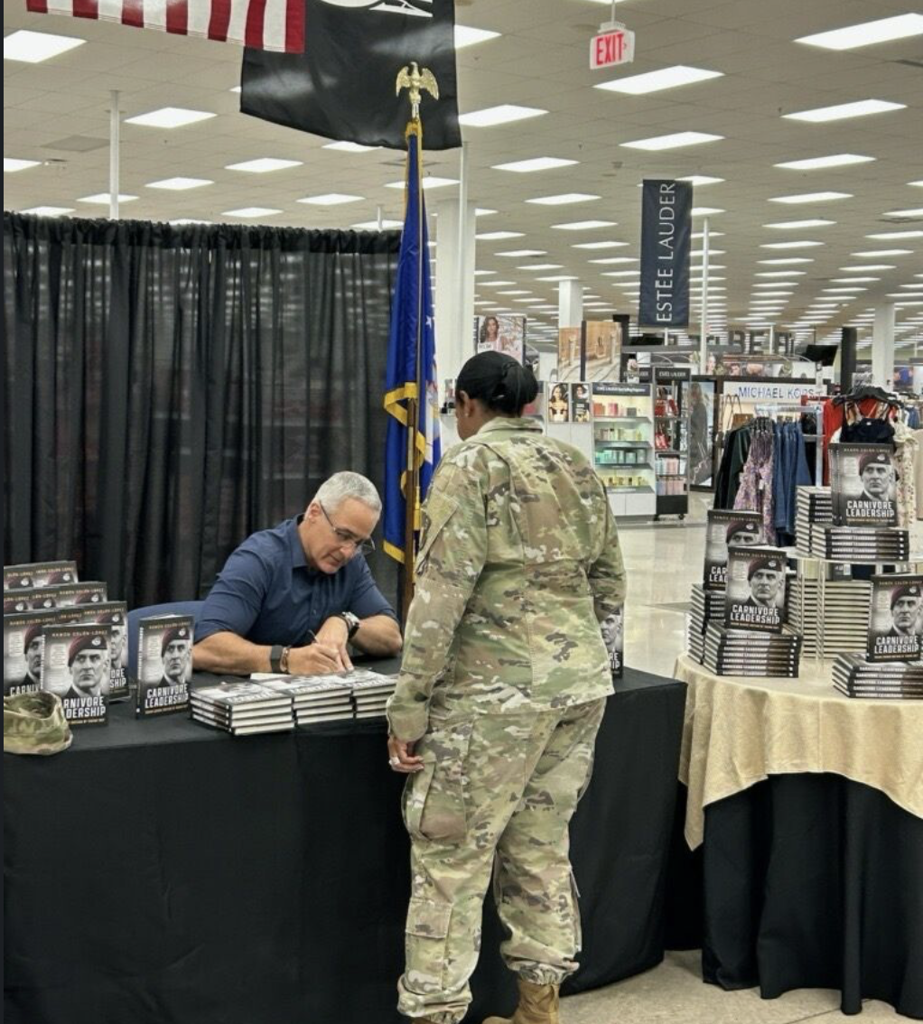 A man in military camouflage signing a book at a table, while a woman in military uniform stands in front of him. The table displays copies of a book titled "Carnivore Leadership." The setting is an indoor store or event space with books, clothing, a