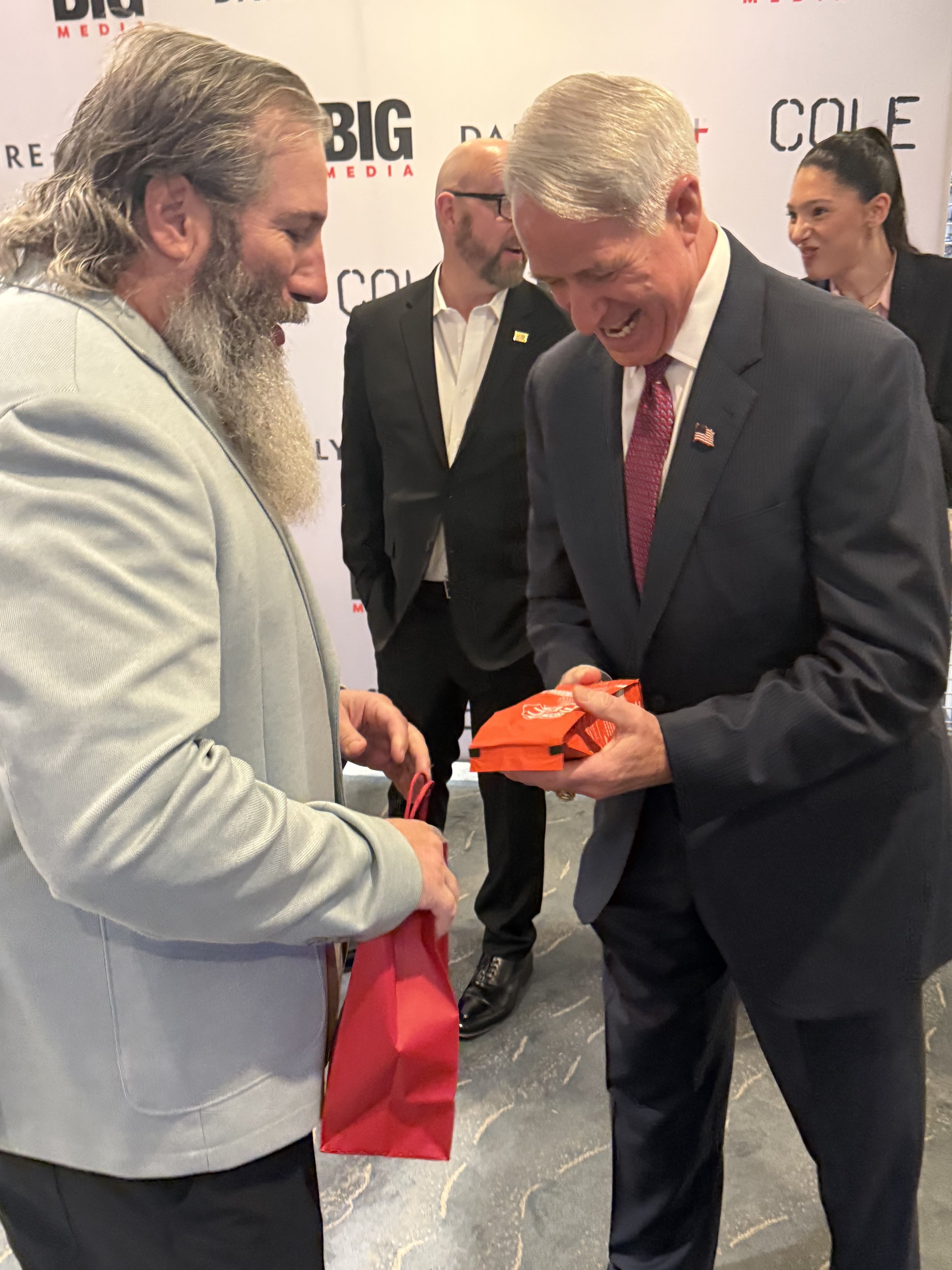 Two men in suits are smiling and shaking hands. One man has long gray hair and a beard, the other has short gray hair. They are at an event with a backdrop that has text including 'BIG', 'MEDIA', and 'COLE'. A woman with dark hair tied back is in the