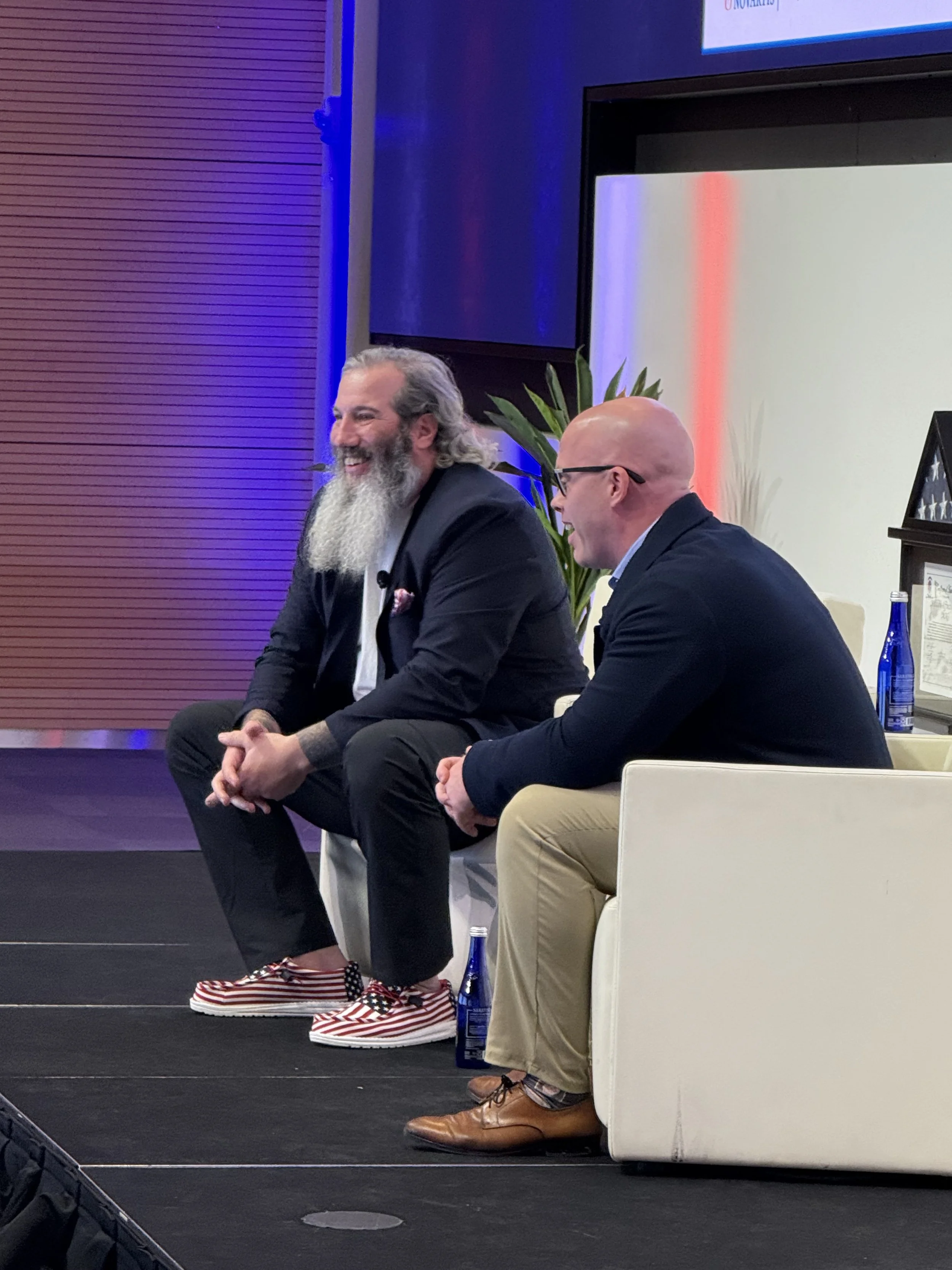 Two men are seated on stage at a panel or event. One man has long gray hair and a beard, wearing a black suit, and the other bald, wearing glasses and a dark blazer. They are smiling and appear engaged in conversation. There are large screens behind 