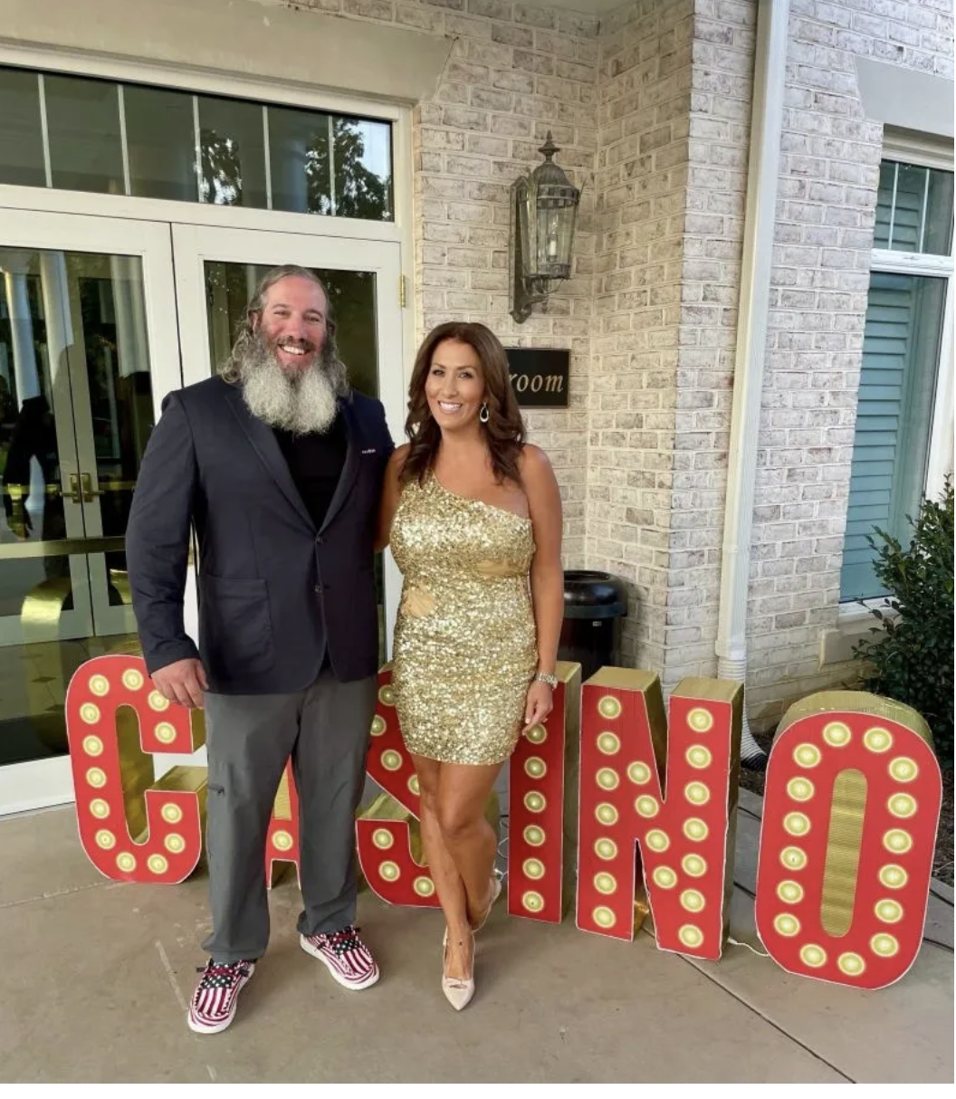 A man with a long gray beard wearing a dark blazer, black shirt, and patterned shoes, standing next to a woman in a gold sequin dress and heels, smiling outdoors near a decorative "casino" sign with large, red marquee letters and bulbs, in front of a