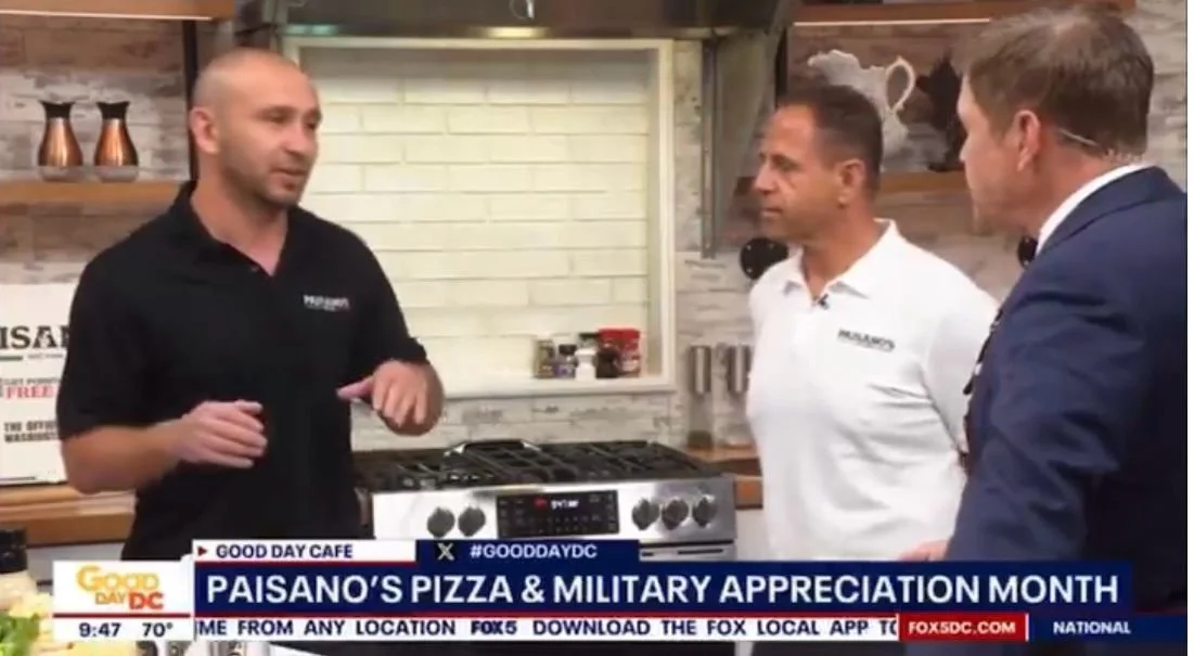 Three men are standing in a kitchen, engaged in a conversation. One man is wearing a black shirt, another is in a white shirt, and the third is in a navy blazer. There is a stove and kitchen shelves behind them.