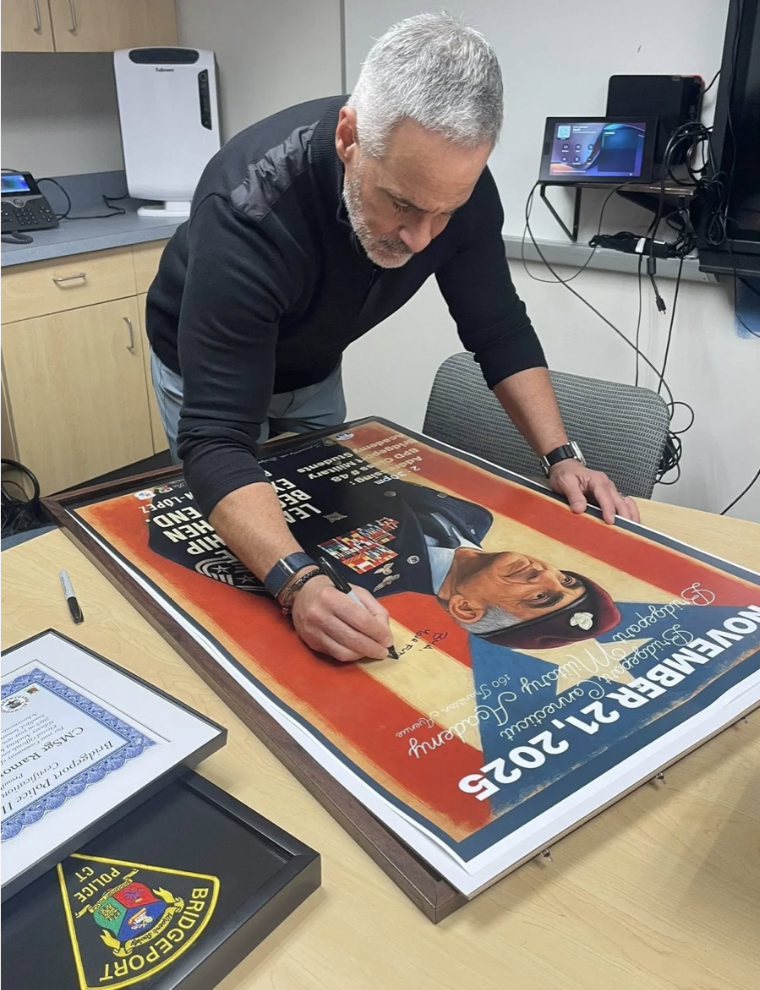 A man with gray hair and a beard, wearing a black shirt, is signing a large framed poster of a person in a military uniform on a table. The background shows a cabinet with a document camera and a television monitor.