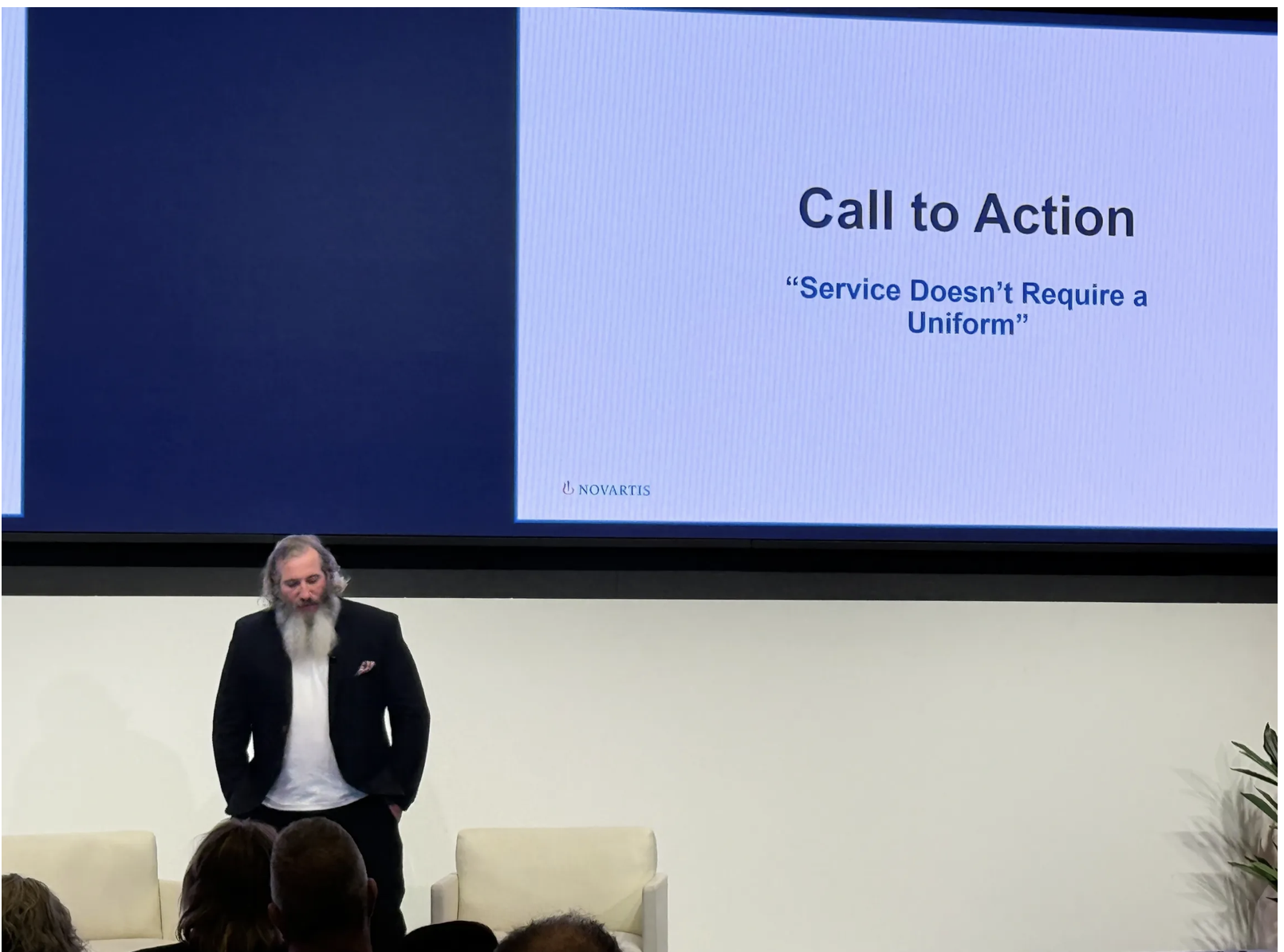 A man with long gray hair and a beard in a black blazer stands in front of a large screen displaying a presentation slide that reads 'Call to Action: Service Doesn't Require a Uniform,' with the Novartis logo at the bottom.