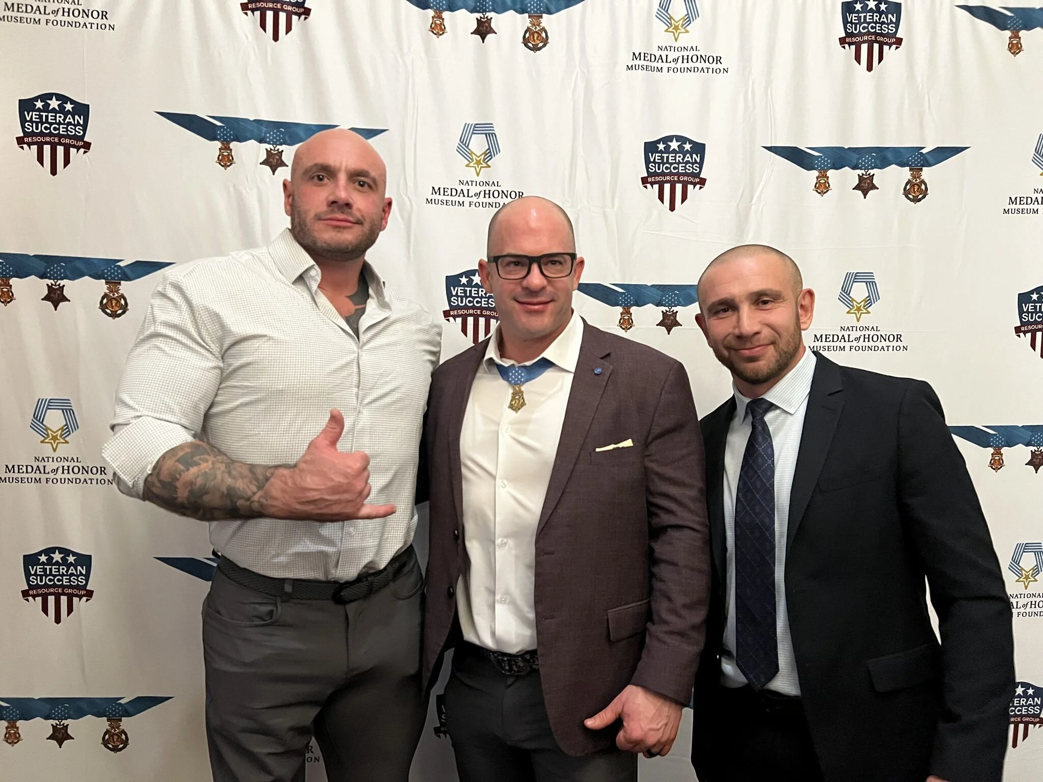 Three men in formal attire standing in front of a backdrop with military medals and banners for the National Medal of Honor Museum Foundation, smiling and posing for a photo.