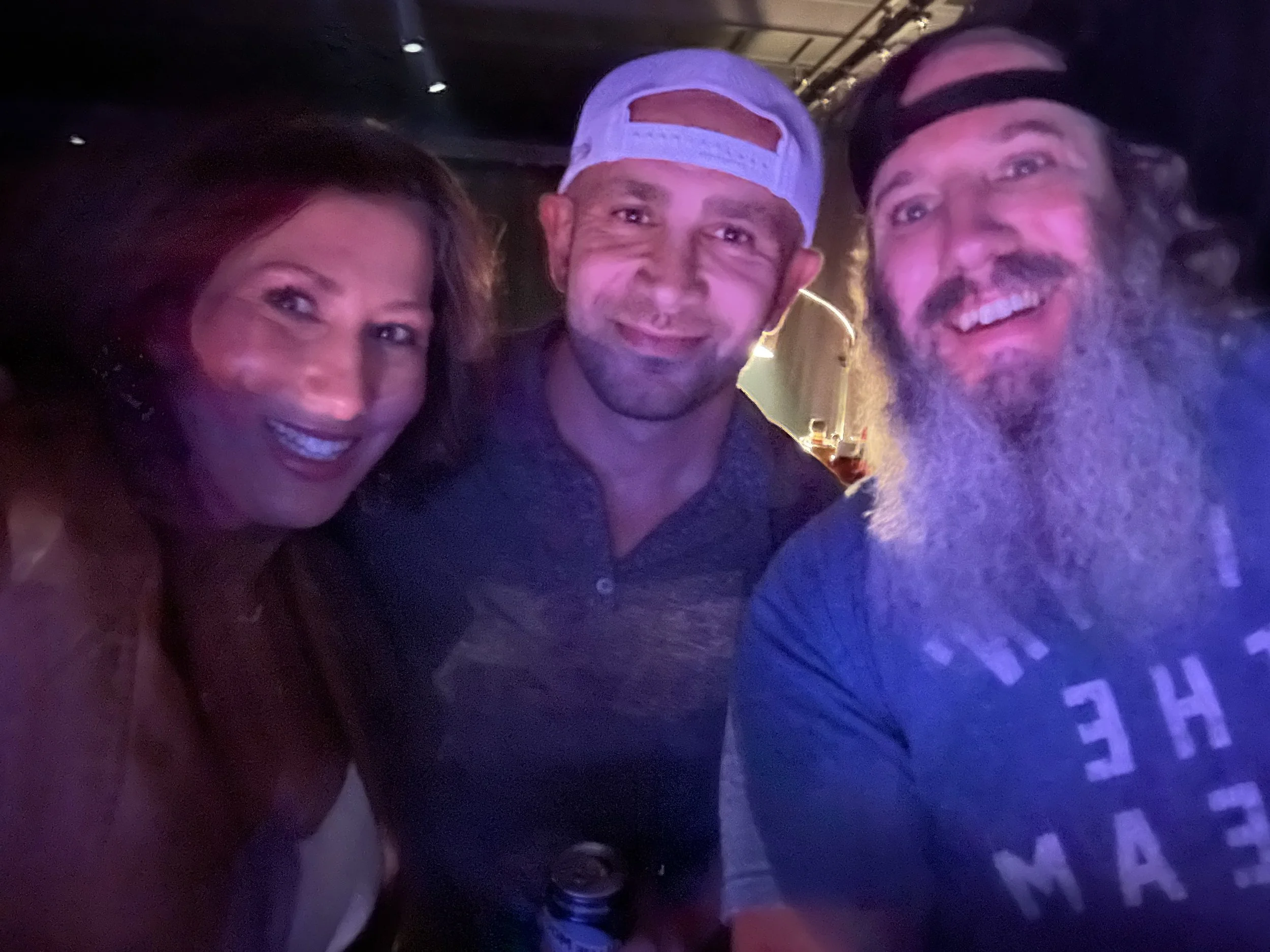 Four friends smiling and taking a group selfie in a dimly lit environment, possibly a bar or club, wearing casual attire including a baseball cap and a backward cap.
