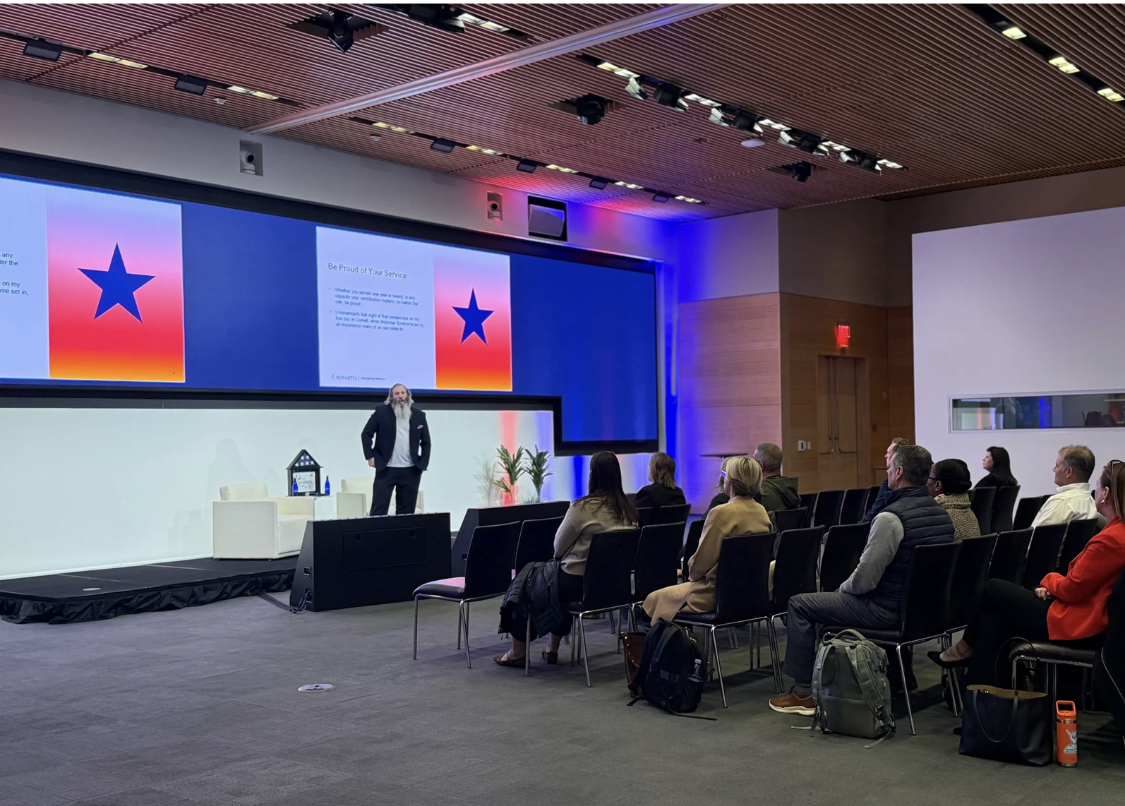 A man with a beard and long hair, dressed in a dark suit, giving a presentation in a conference room with an audience seated facing him. The large screen behind displays a blue star on a gradient background with pink, orange, and blue colors. The roo