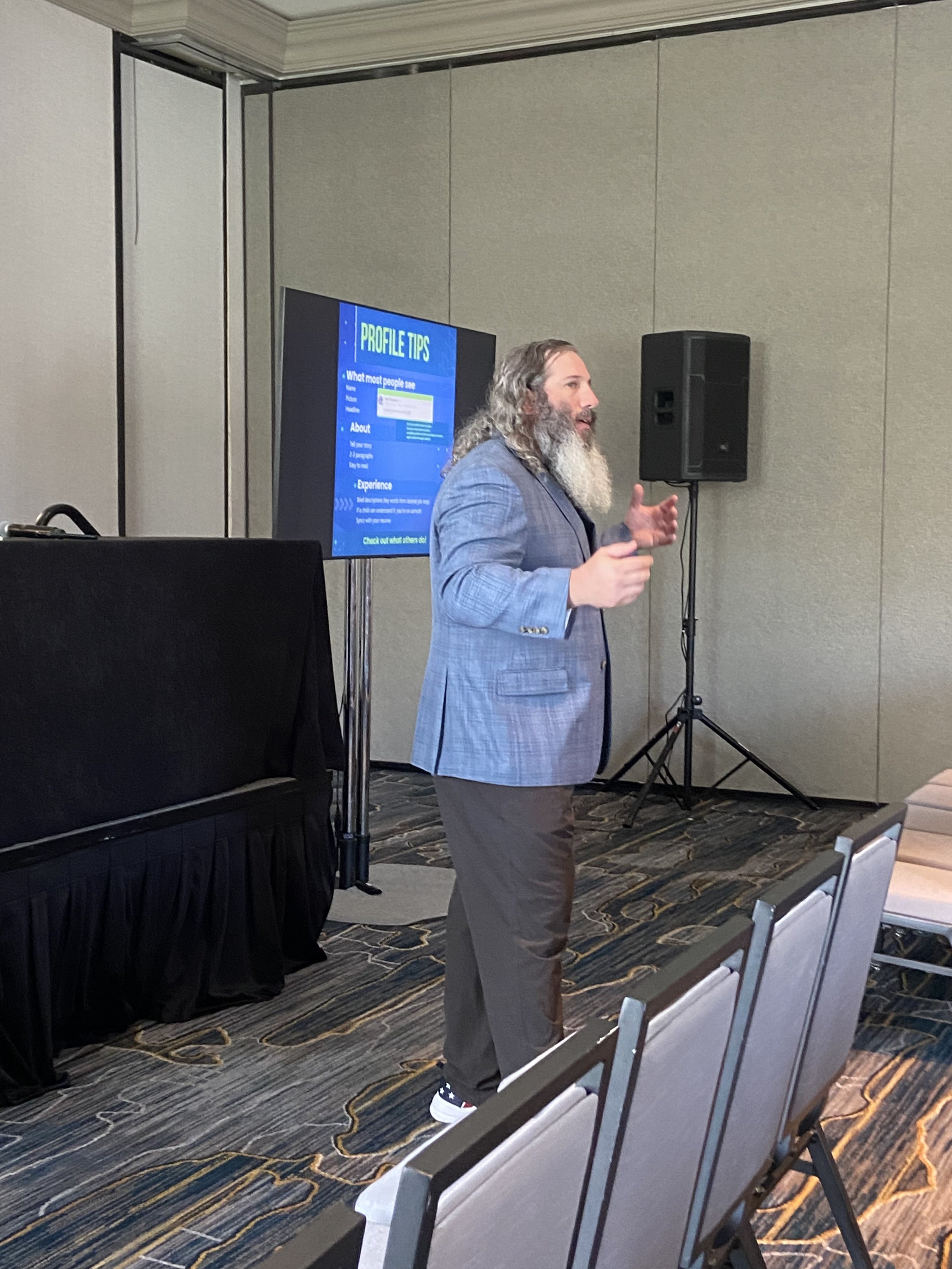 A man with long hair, a beard, wearing a light blue plaid blazer and gray pants, speaking at a conference or presentation in a room with a projector screen displaying "Profile Tips" and a black speaker on a stand.