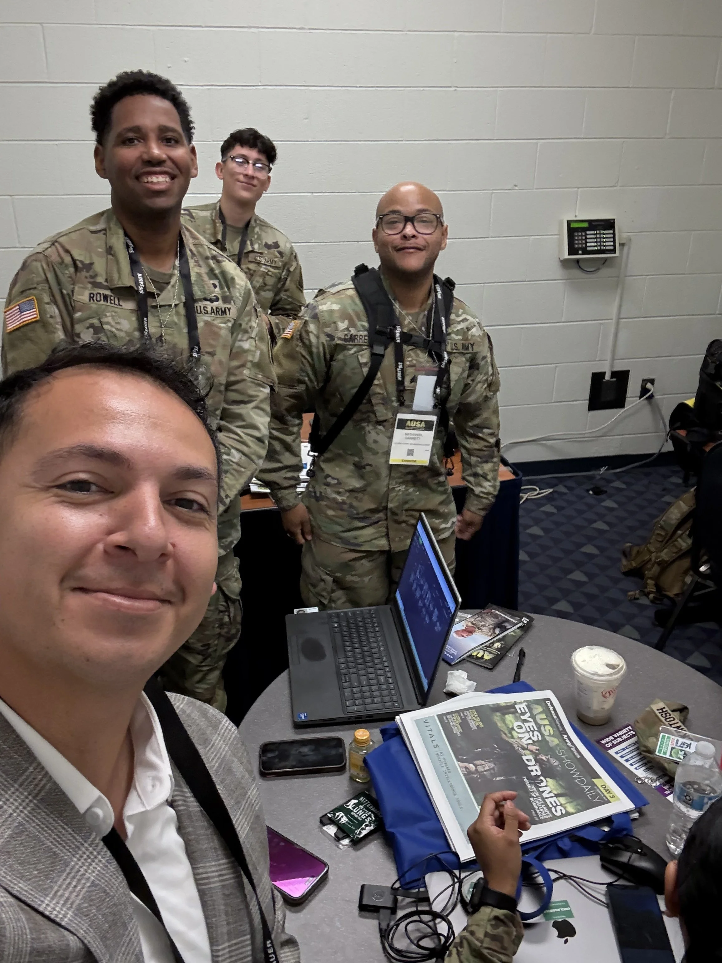 Four men in military fatigues gathered around a table at a conference or event, smiling at the camera. The table has laptops, magazines, water bottles, and other items on it. A gray wall and some electronic equipment are in the background.