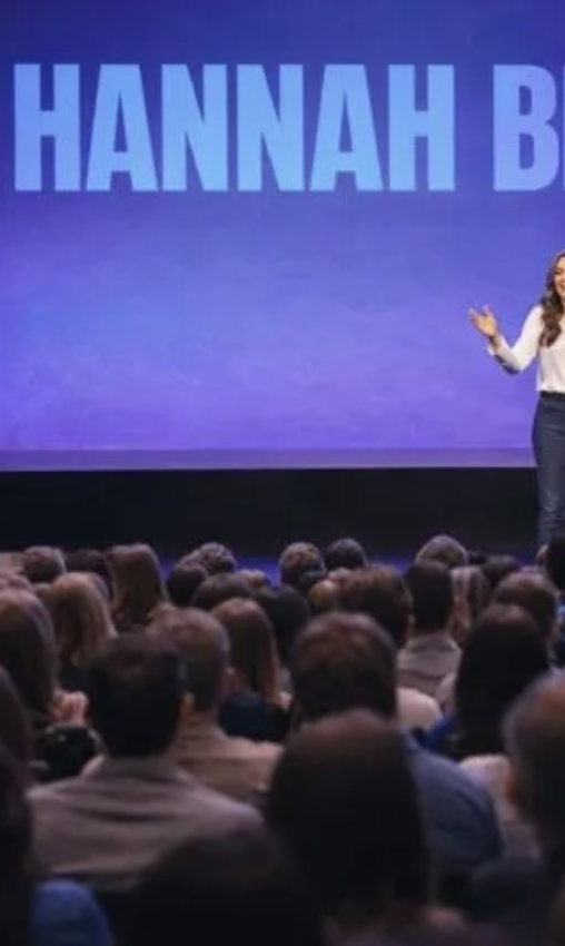 A woman standing on stage speaking to an audience, with the name "Hannah B" displayed on a large screen behind her.