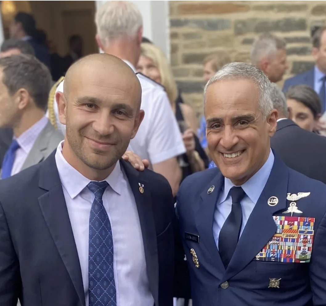 Two men in military uniforms at a social event, smiling for the camera. The man on the right is wearing a decorated uniform with medals and insignia, and the man on the left is wearing a suit and tie. Other people are in the background.