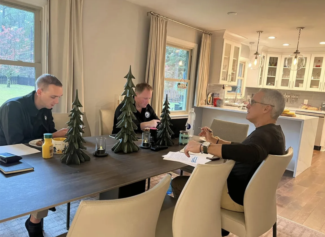 Three people sitting at a dining table in a kitchen, three decorative trees in the center, with windows showing an outdoor yard and decorated for the holidays.