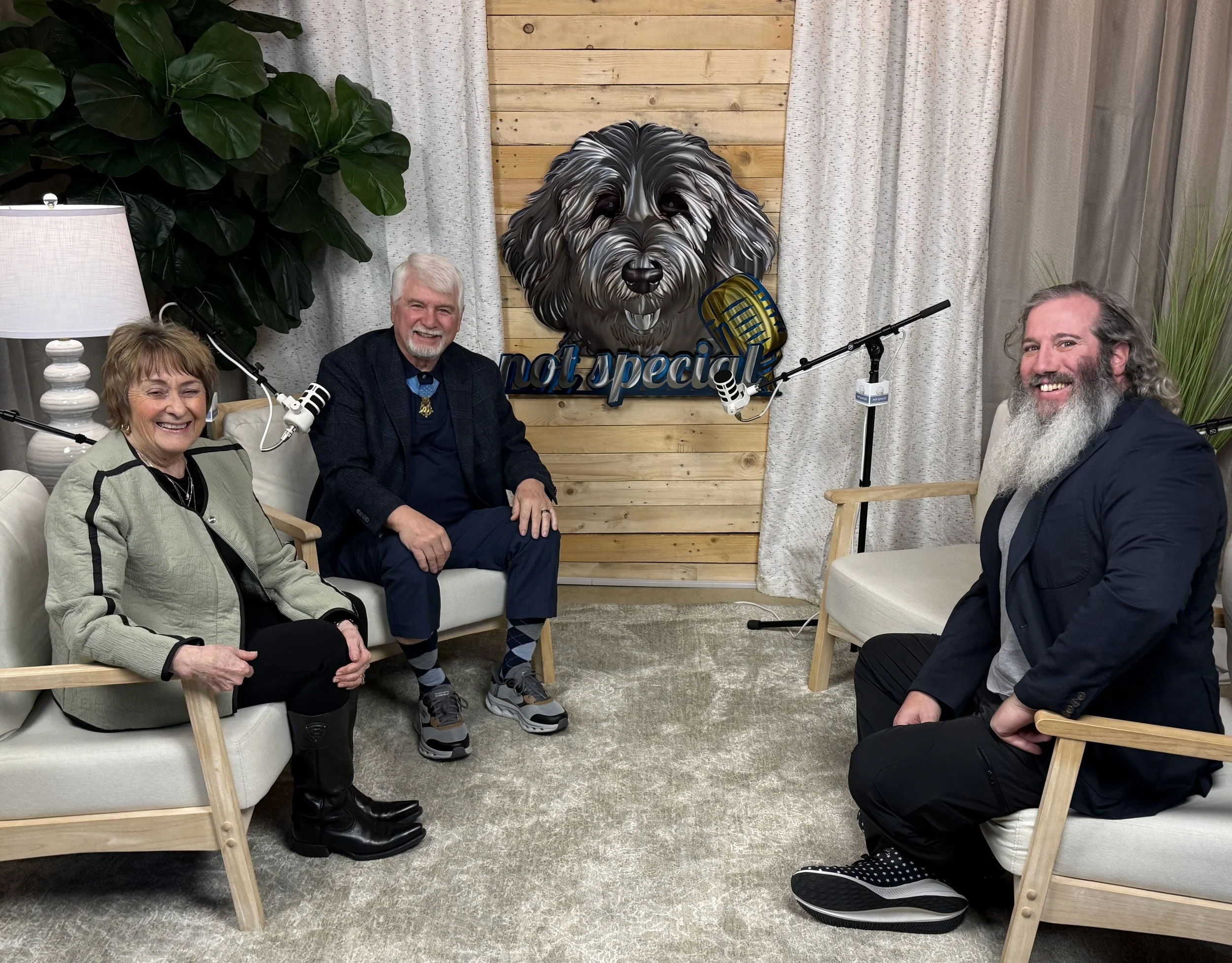 Three people are sitting in chairs inside a recording studio, smiling at the camera. There is a large, colorful wall art of a dog with a microphone in front of it and the text 'not special' underneath. The background features a wooden wall panel, cur