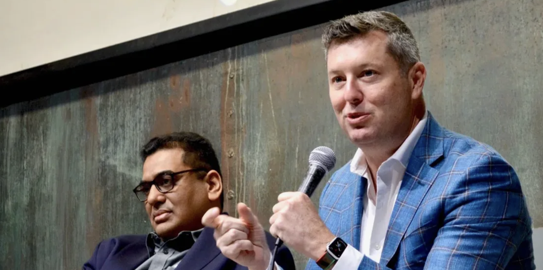 Two men sitting at a panel discussion, one wearing glasses and a navy suit, the other speaking into a microphone while wearing a blue plaid blazer, white shirt, and smartwatch, with a textured wall behind them.