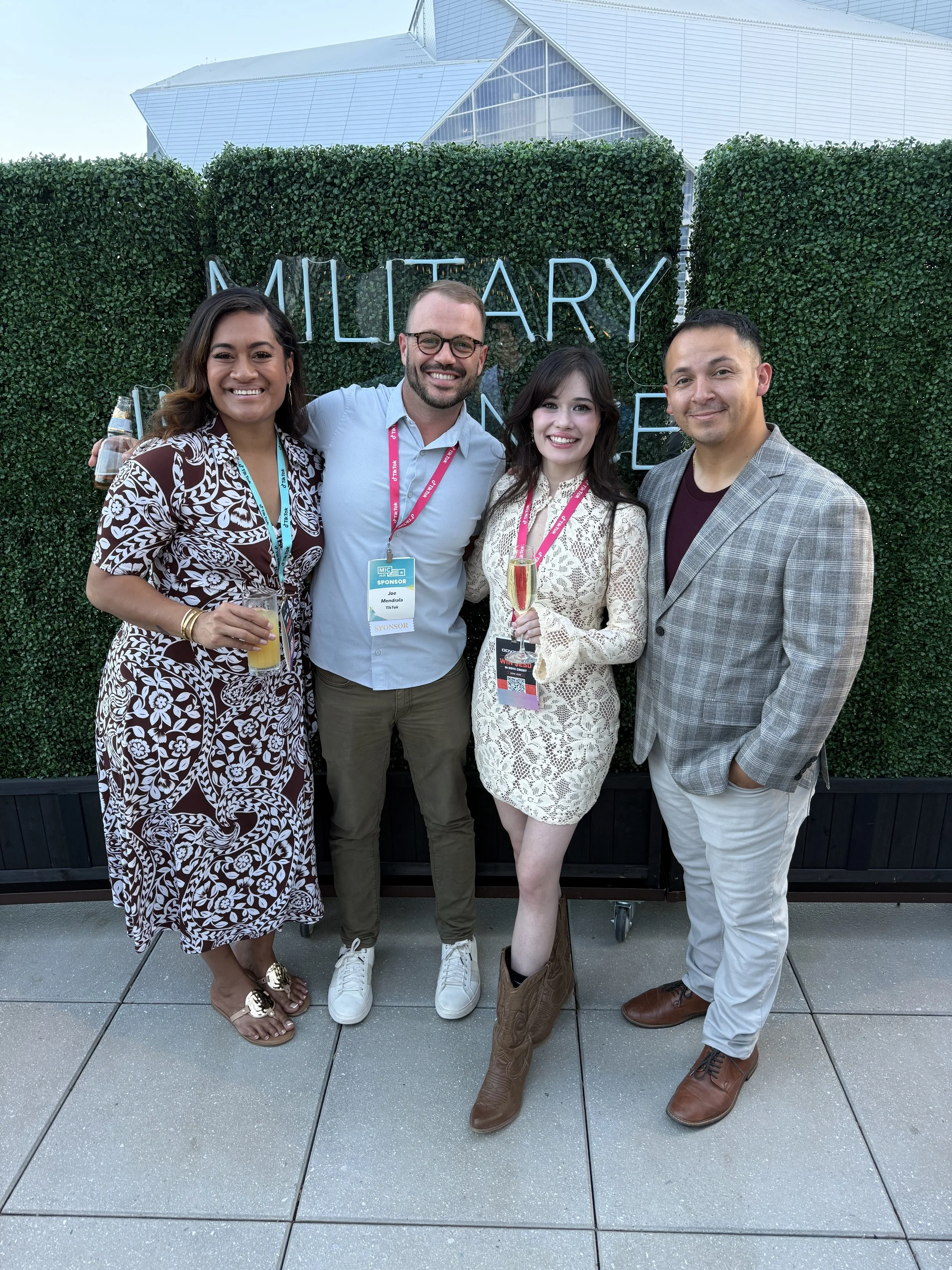 Four people standing together in front of a green hedge with a illuminated sign that reads 'MULTIARY' in the background. Two women and two men, dressed in business casual attire, holding drinks, posing for a photo outdoors on a tiled surface.