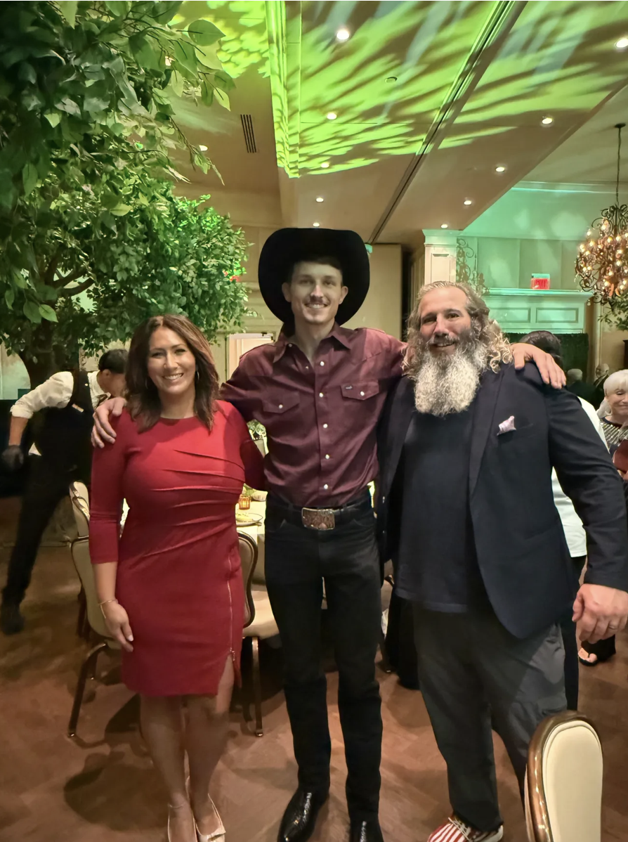 Three people smiling and posing together at a festive event indoors. The woman on the left is wearing a red dress, the man in the middle is wearing a maroon shirt and black hat, and the man on the right has a beard and curly hair, wearing a black bla
