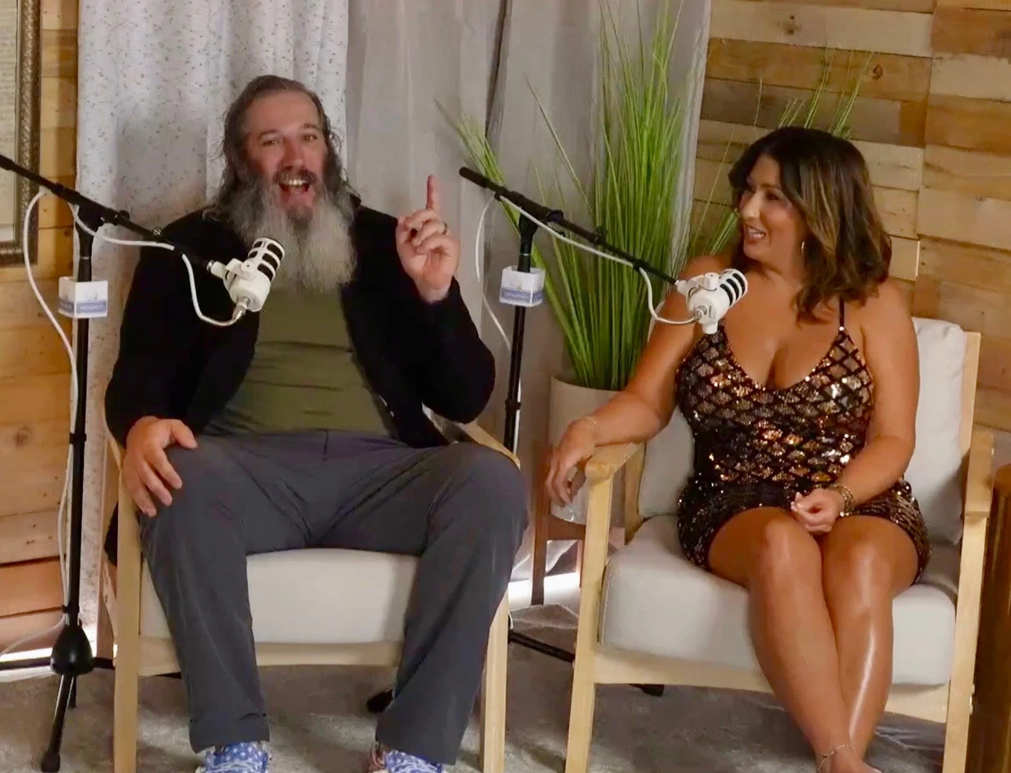 A man with a long gray beard and a woman with shoulder-length wavy brown hair are sitting together on armchairs, engaging in conversation. They are in a cozy room with wooden walls, a large green plant, and microphones set up in front of them, sugges