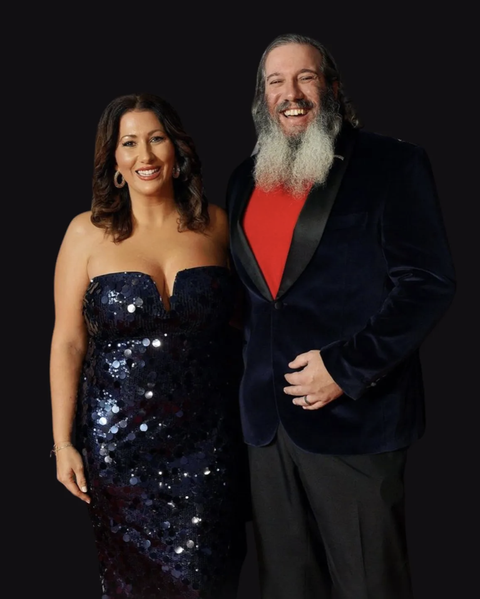 A woman in a strapless, dark blue, sequined gown and a man in a navy blazer with a red shirt and black pants, both smiling against a black background.