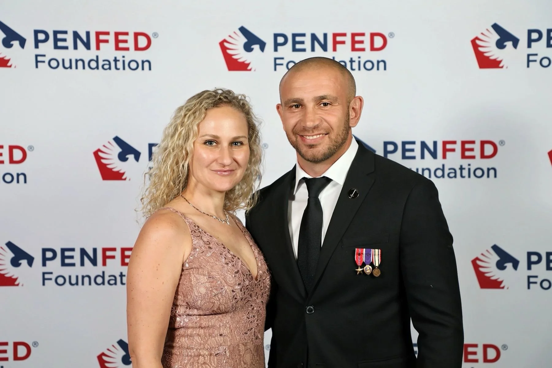 Man and woman standing together at PenFed Foundation event, woman in pink lace dress, man in military uniform with medals, background with PenFed Foundation logos.