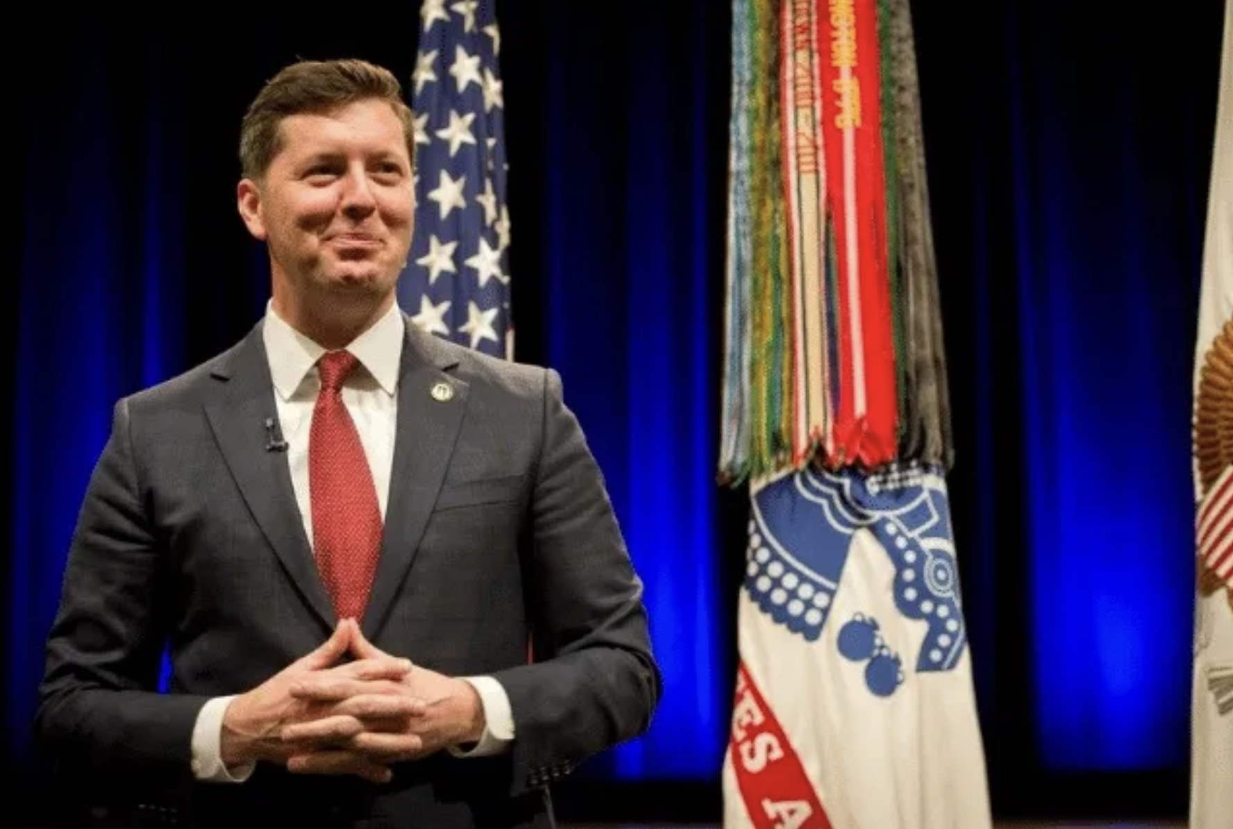 A man in a gray suit with a red tie standing on a stage with blue curtains and flags, including an American flag and various banners, in the background.