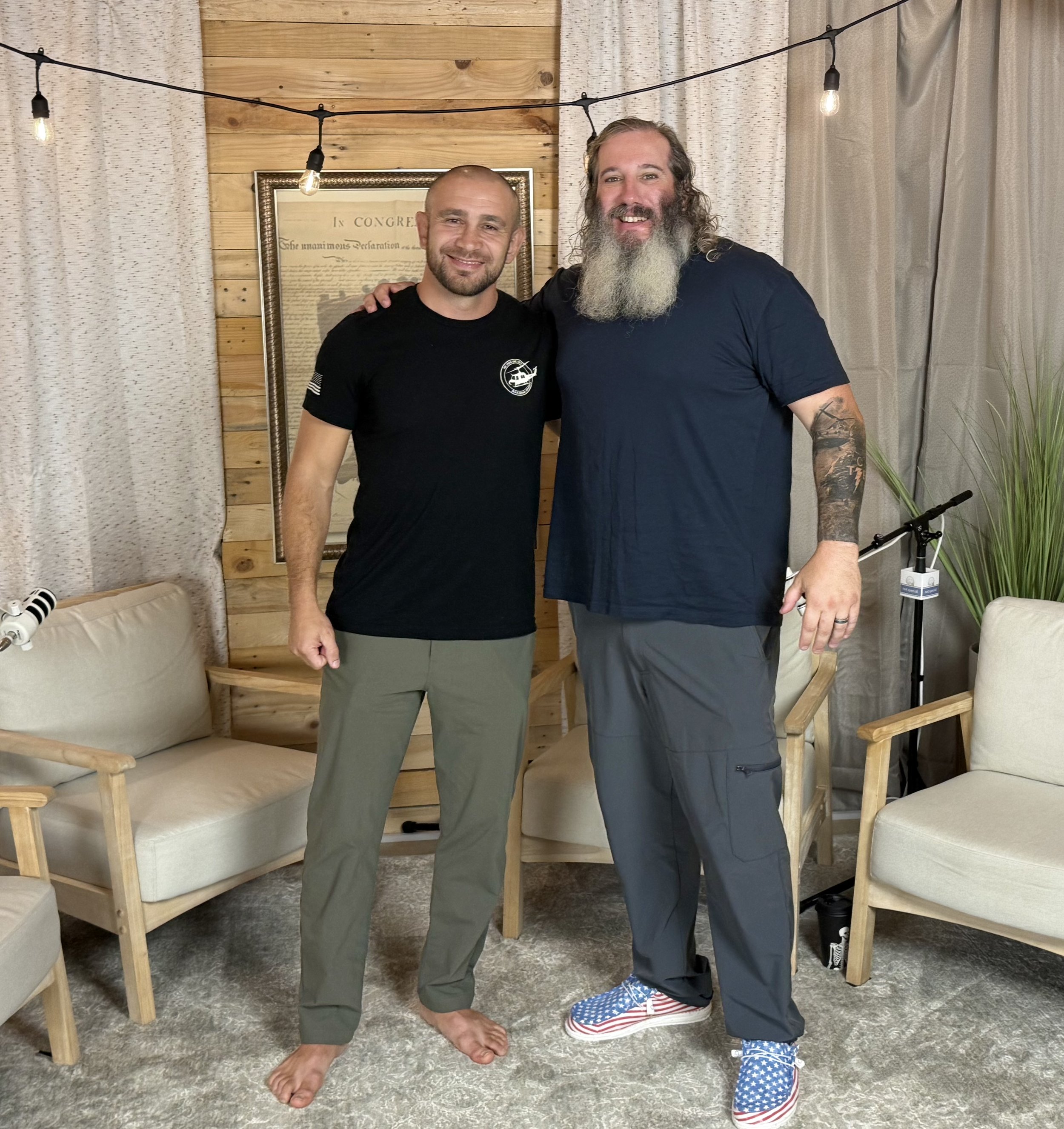 Two men standing indoors with their arms around each other, smiling at the camera. The man on the left is wearing a black t-shirt and beige pants, and the man on the right is wearing a dark t-shirt and dark pants, with comic-style sneakers. The backg