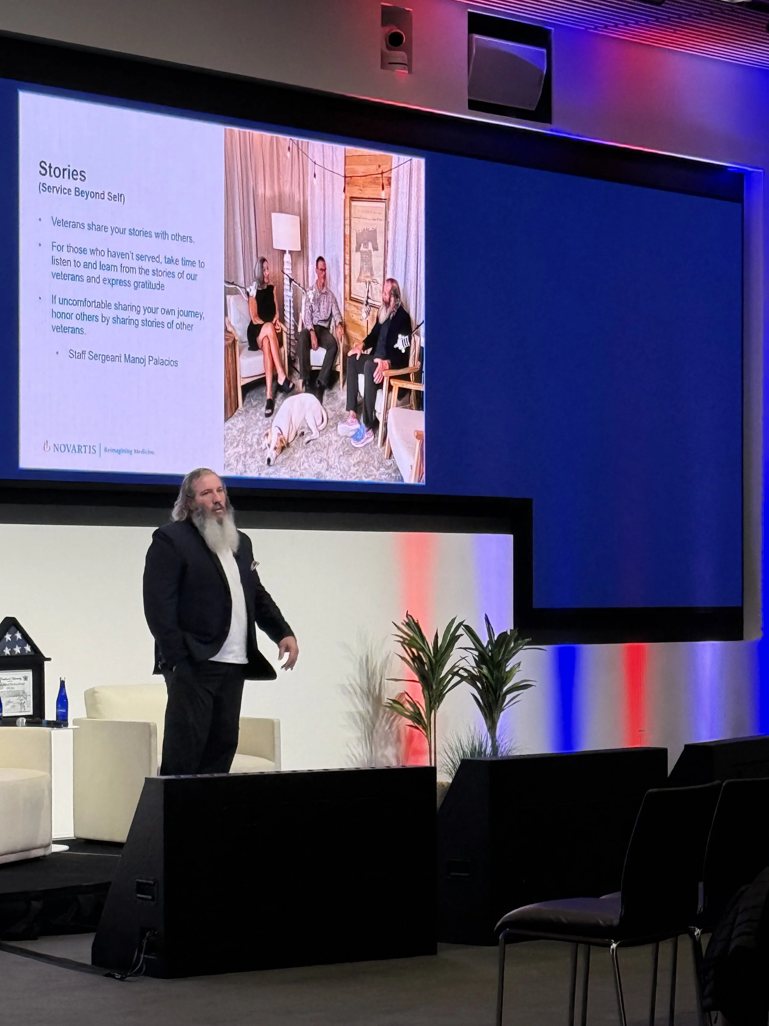 A man with long gray hair and a beard is standing on a stage in a conference room. Behind him, there are chairs, plants, and a large screen displaying a slide titled "Stories" with text and a photo of three people sitting in a room. The environment i