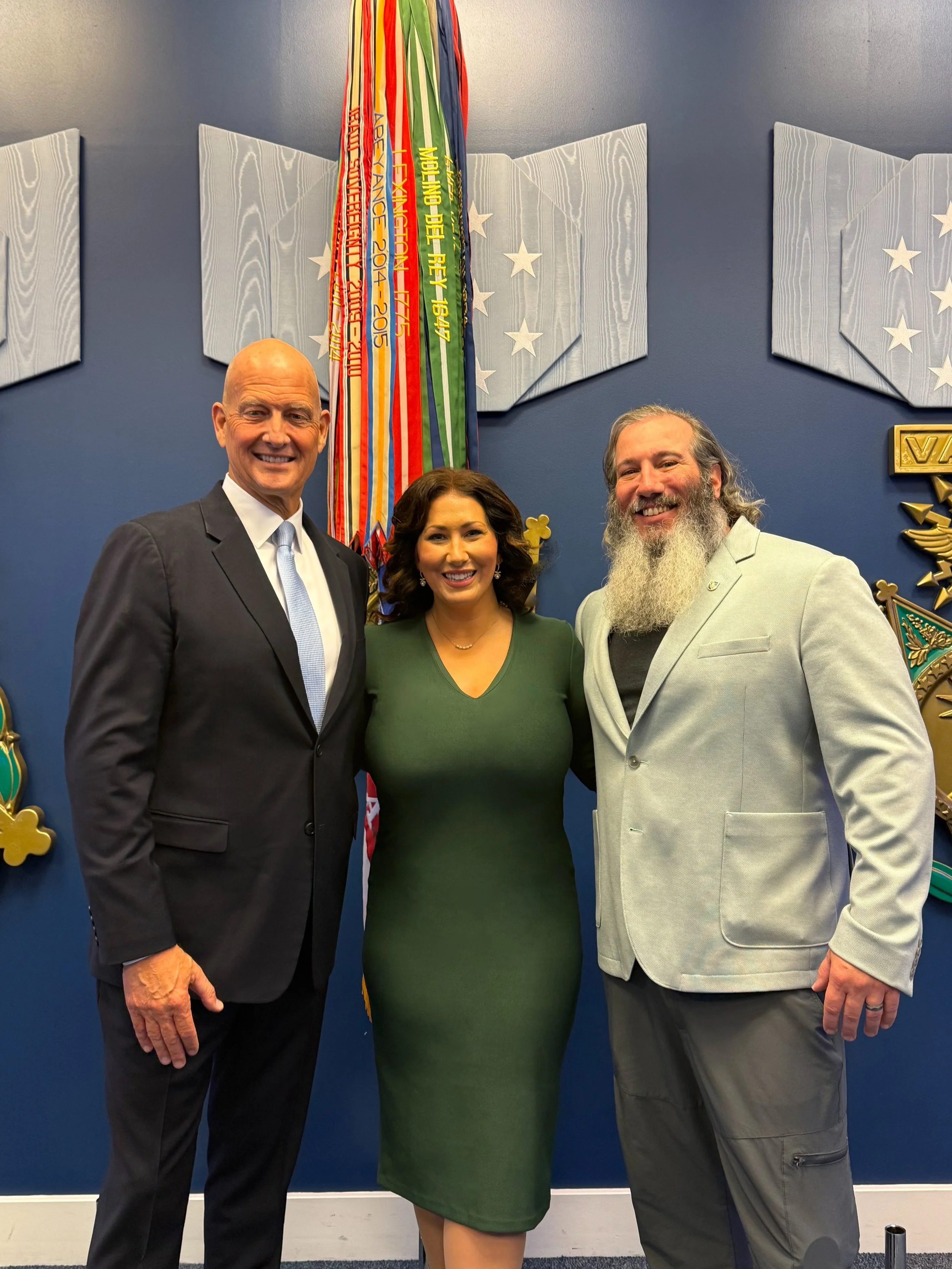 Three people, two men and one woman, standing together smiling in front of a blue wall with medals and banners hanging on it.