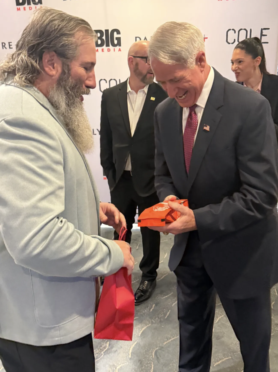 Two men, one with long gray hair and a beard wearing a light gray suit, and the other with short gray hair in a dark suit and red tie, are smiling and exchanging gifts at an event. There are two other people in the background, a man with glasses and 
