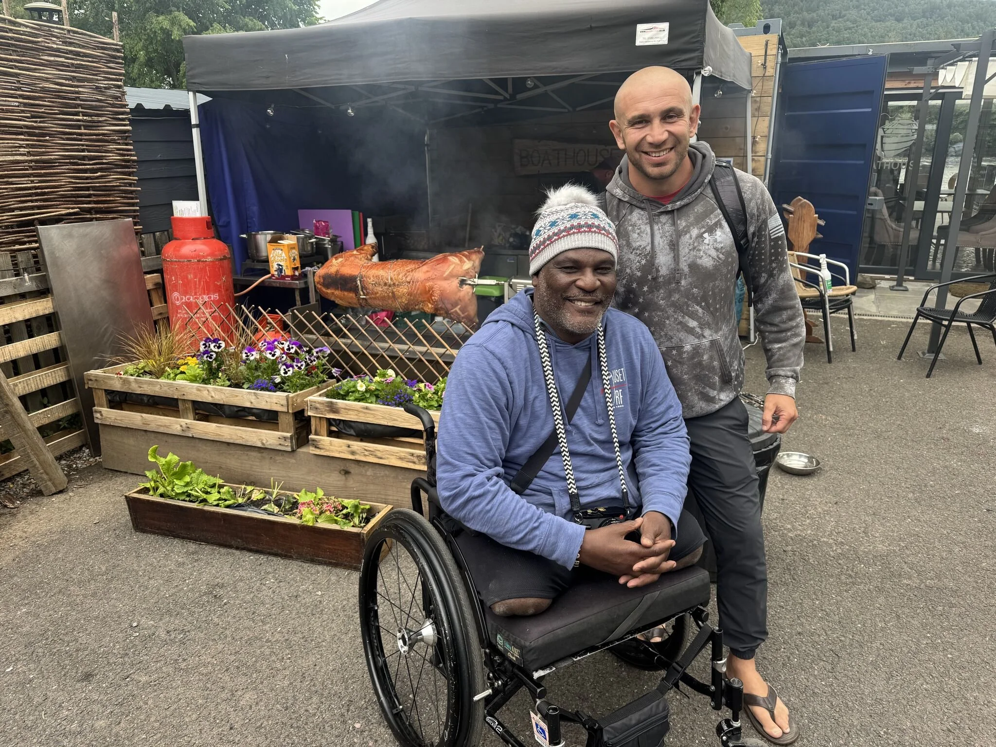 Two smiling men, one in a wheelchair, in an outdoor area with flowers and a barbecue grill behind them. One man is bald, wearing a gray hoodie, and standing; the other man has a beard, wearing a gray and white cap, and sitting in the wheelchair.