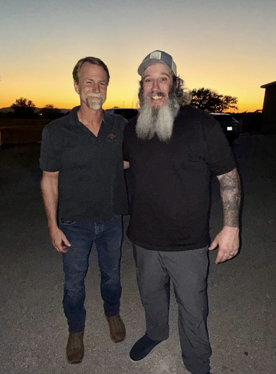Two men standing outdoors at sunset, smiling, with trees and a building in the background. One man has a beard and is wearing a baseball cap and dark clothing; the other has a beard and is wearing a dark polo shirt and jeans.