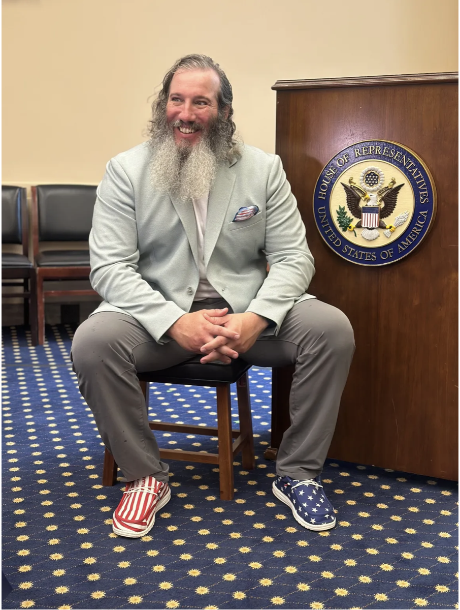 A man with long gray hair and beard wearing a light gray suit, sitting on a chair, smiling, in a room with a blue and yellow patterned carpet, beside a wooden podium with the seal of the United States House of Representatives. He is wearing patriotic